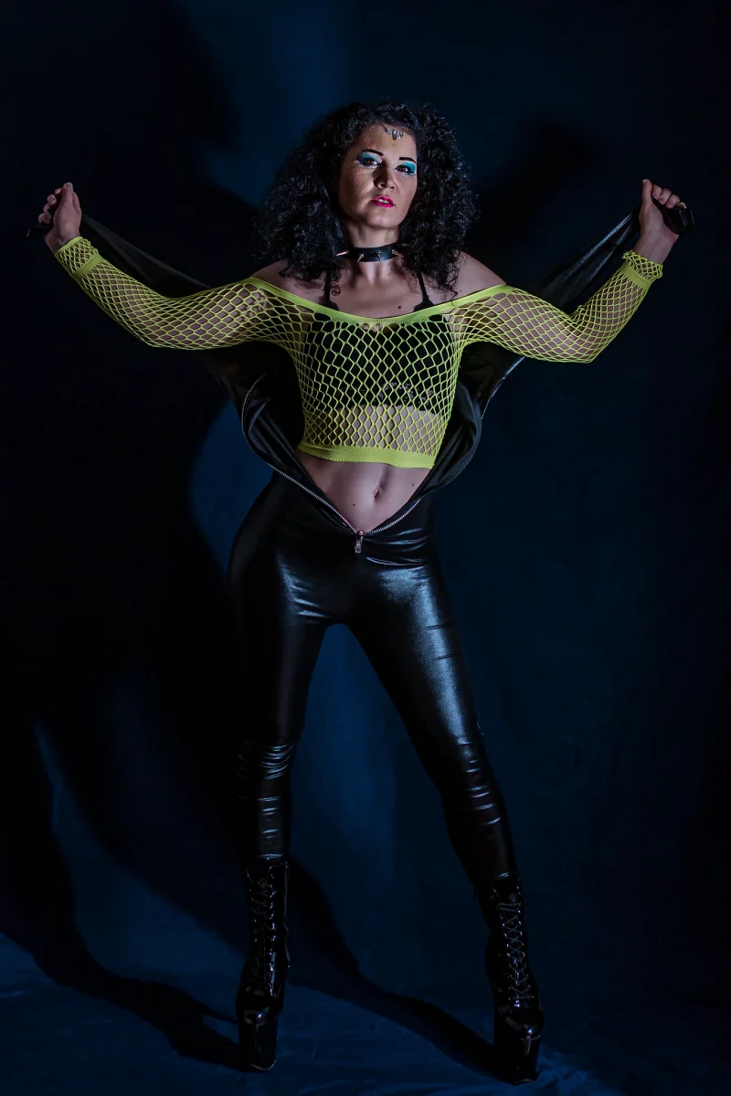 Creative - Porträt einer Frau in dunkler Studioszene, die selbstbewusst vor einem schwarzen Hintergrund steht; sie trägt einen geöffneten schwarzen Catsuit, dessen Oberteil hinter ihrem Rücken gespannt ist und ihren Oberkörper freilässt, kombiniert mit einem neonfarbenen Netzoberteil und hohen Stiefeln. Mit ausgebreiteten Armen hält sie den Catsuit straff, während gerichtetes Licht starke Kontraste erzeugt und die Szene eine markante, edgy Bildwirkung erhält.