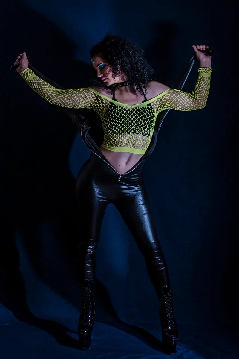 Creative - Porträt einer Frau in dunkler Studioszene, die selbstbewusst vor einem schwarzen Hintergrund steht; sie trägt einen geöffneten schwarzen Catsuit, dessen Oberteil hinter ihrem Rücken gespannt ist und ihren Oberkörper freilässt, kombiniert mit einem neonfarbenen Netzoberteil und hohen Stiefeln. Mit ausgebreiteten Armen hält sie den Catsuit straff, hat den Kopf nach nach rechts unten gedreht, während gerichtetes Licht starke Kontraste erzeugt und die Szene eine markante, edgy Bildwirkung erhält.