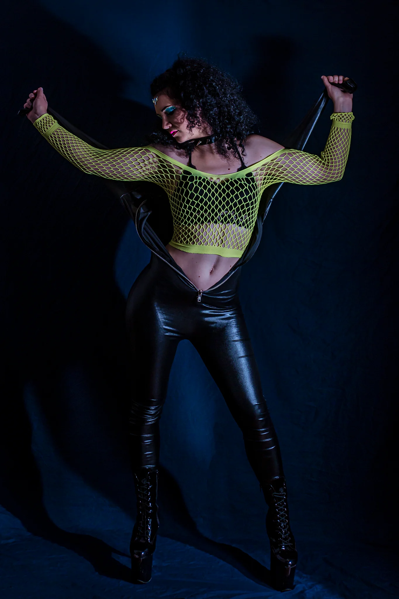 Creative - Porträt einer Frau in dunkler Studioszene, die selbstbewusst vor einem schwarzen Hintergrund steht; sie trägt einen geöffneten schwarzen Catsuit, dessen Oberteil hinter ihrem Rücken gespannt ist und ihren Oberkörper freilässt, kombiniert mit einem neonfarbenen Netzoberteil und hohen Stiefeln. Mit ausgebreiteten Armen hält sie den Catsuit straff, hat den Kopf nach nach rechts unten gedreht, während gerichtetes Licht starke Kontraste erzeugt und die Szene eine markante, edgy Bildwirkung erhält.