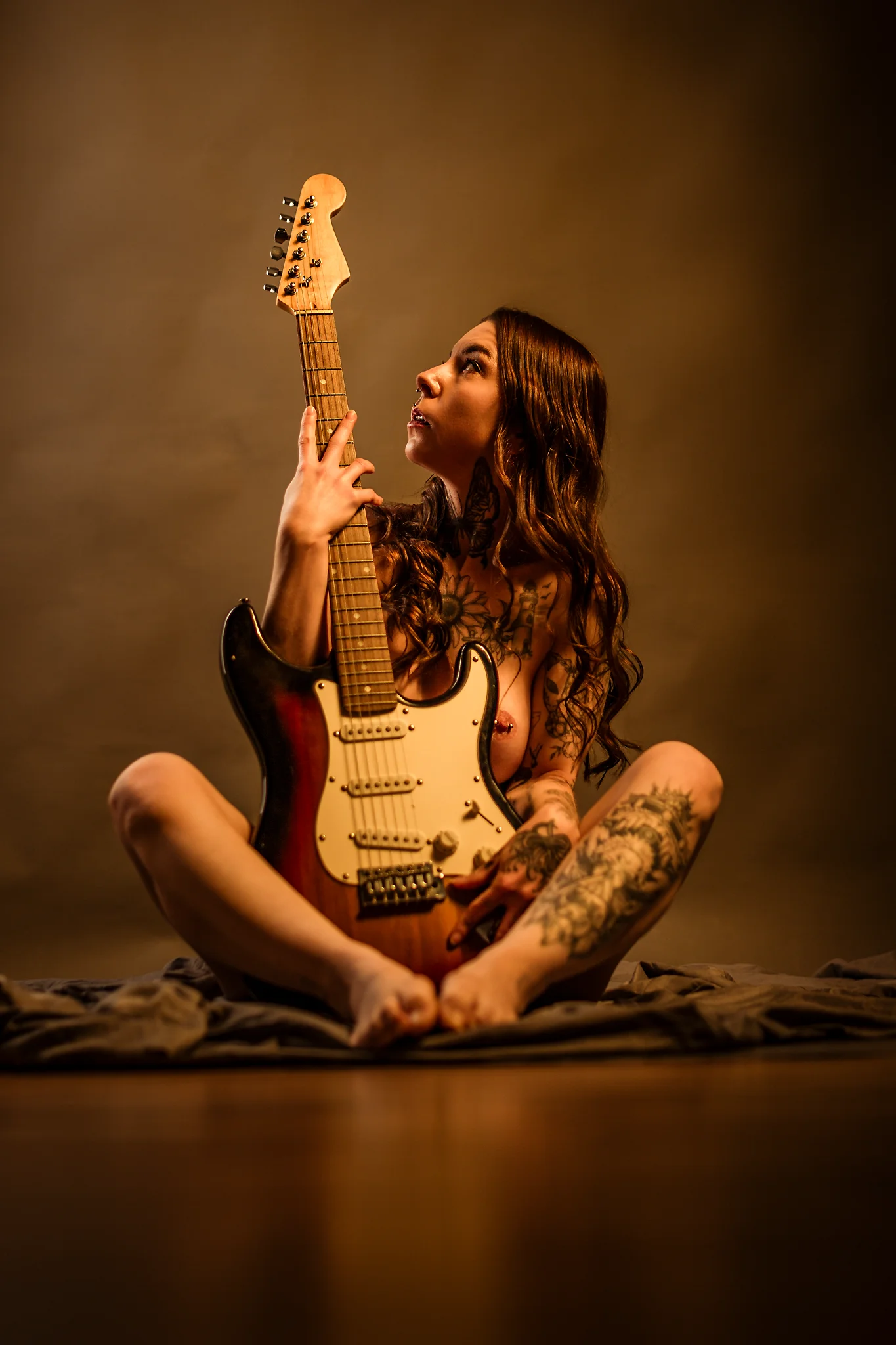 Creative - Sinnliches Porträt einer tätowierten Frau in warmem, weichem Licht, die nackt auf dem Boden sitzt und eine E-Gitarre vor ihrem Körper hält, sodass sie ihren Oberkörper teilweise verdeckt. Sie blickt nach oben entlang des Gitarrenhalses, während die warme Lichtstimmung und der reduzierte Hintergrund eine ruhige, intime Atmosphäre mit künstlerischem Fokus auf Form, Haltung und Instrument erzeugen.