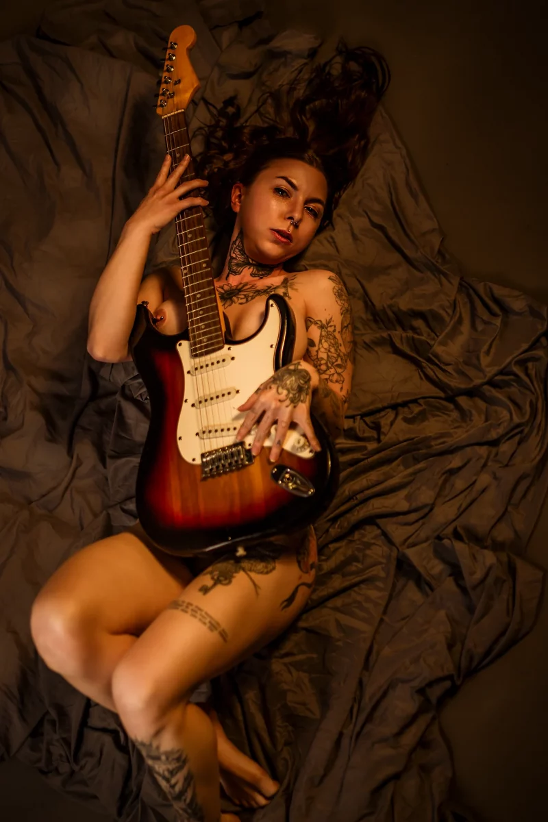 Creative - Sinnliches Porträt einer tätowierten Frau in warmer, weicher Lichtstimmung, die nackt auf zerknittertem Stoff liegt und eine E-Gitarre eng an ihren Körper zieht, sodass ihr Oberkörper teilweise bedeckt ist. Ihr Blick wirkt ruhig und direkt, während das warme Licht Haut, Tattoos und die glänzende Oberfläche des Instruments hervorhebt und eine intime, künstlerische Atmosphäre schafft.