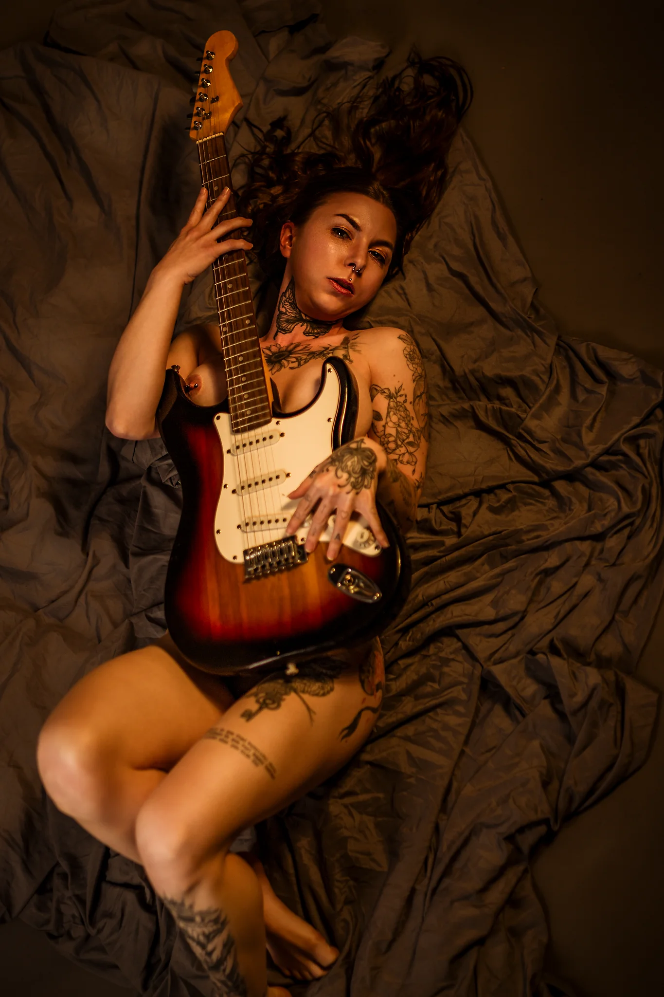 Creative - Sinnliches Porträt einer tätowierten Frau in warmer, weicher Lichtstimmung, die nackt auf zerknittertem Stoff liegt und eine E-Gitarre eng an ihren Körper zieht, sodass ihr Oberkörper teilweise bedeckt ist. Ihr Blick wirkt ruhig und direkt, während das warme Licht Haut, Tattoos und die glänzende Oberfläche des Instruments hervorhebt und eine intime, künstlerische Atmosphäre schafft.