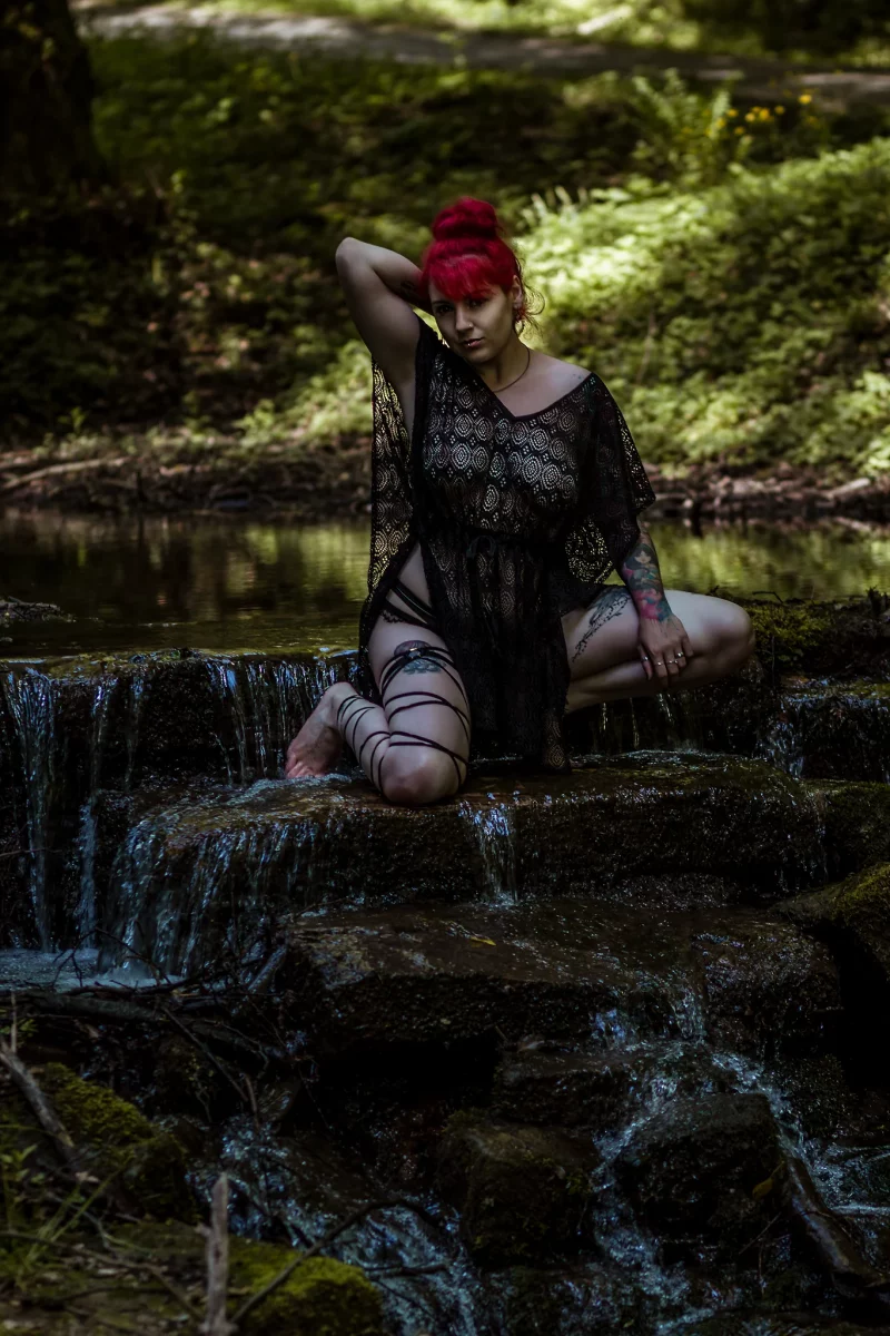 Natur - Person mit roten Haaren sitzt auf einer kleinen Felskaskade in einem Bachlauf im Wald, trägt ein transparentes schwarzes Kleid mit Dessous darunter und blickt selbstbewusst in die Kamera; stimmungsvolle Naturaufnahme mit weichem Licht und fließendem Wasser.