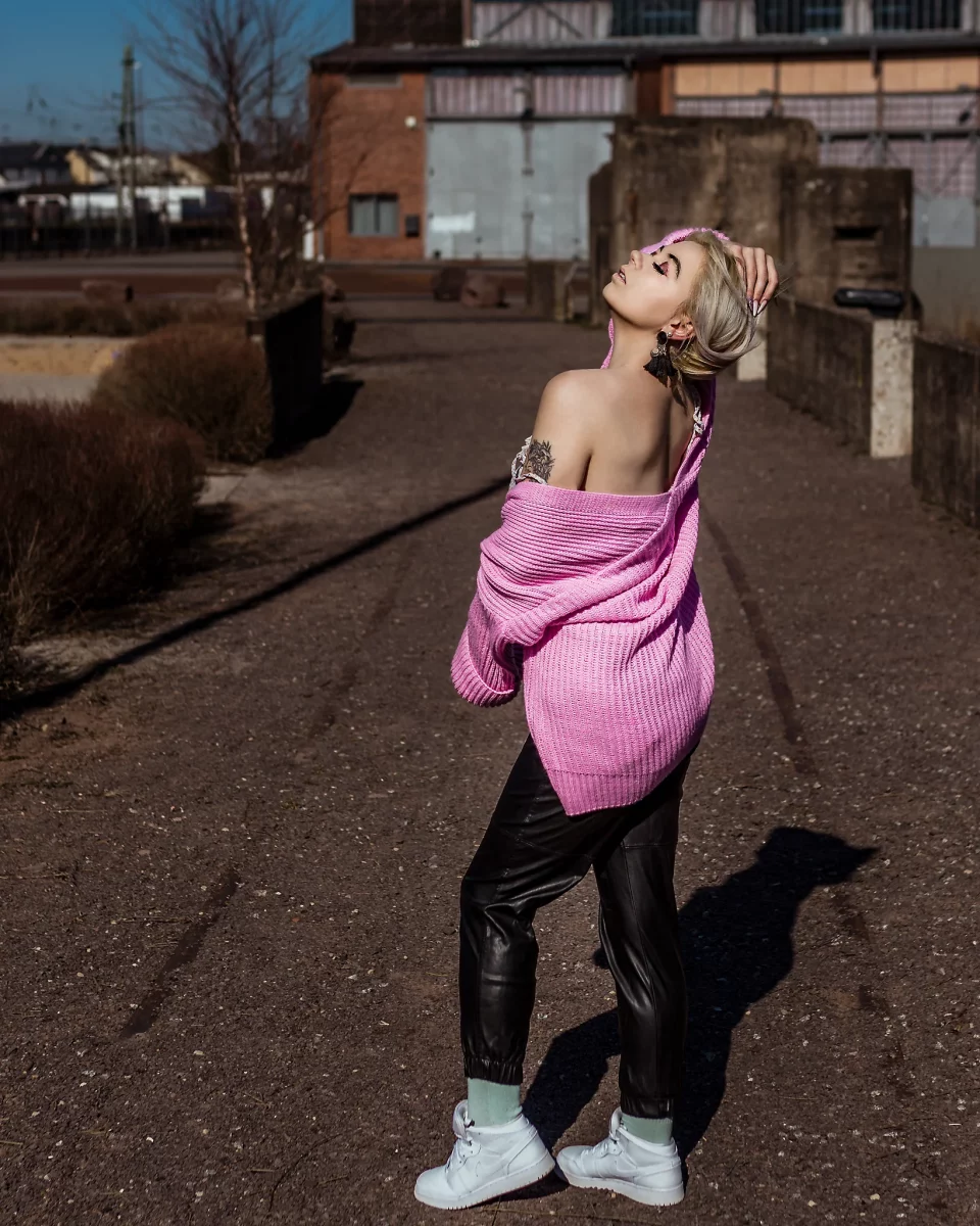 City - Person steht in einer urbanen Umgebung vor industrieller Architektur, trägt eine locker fallende Strickjacke und blickt mit nach hinten geneigtem Kopf in das Licht. Die Pose wirkt selbstbewusst und offen, während hartes Sonnenlicht klare Schatten und Konturen erzeugt. Die Kombination aus rauer Umgebung und farblichem Kontrast schafft eine moderne, leicht edgy Bildwirkung.