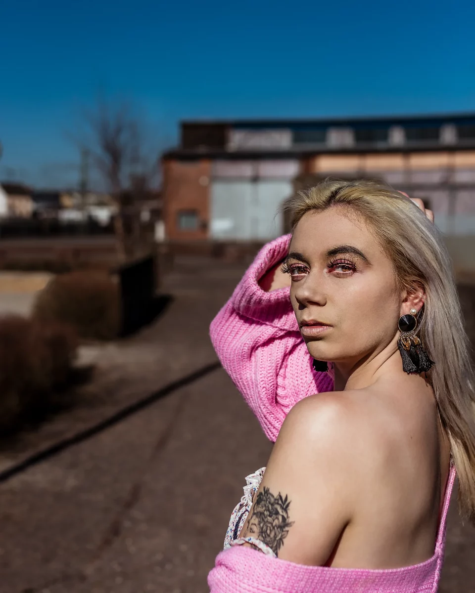 City - Person steht in einer urbanen Umgebung vor industrieller Architektur, die Strickjacke locker über die Schultern gelegt und mit direktem, fokussiertem Blick über die Schulter zur Kamera. Hartes Sonnenlicht erzeugt klare Schatten und betont Haut sowie Konturen, während der unscharfe Hintergrund und die kräftigen Farben eine moderne, kontrastreiche und leicht edgy Bildwirkung schaffen.