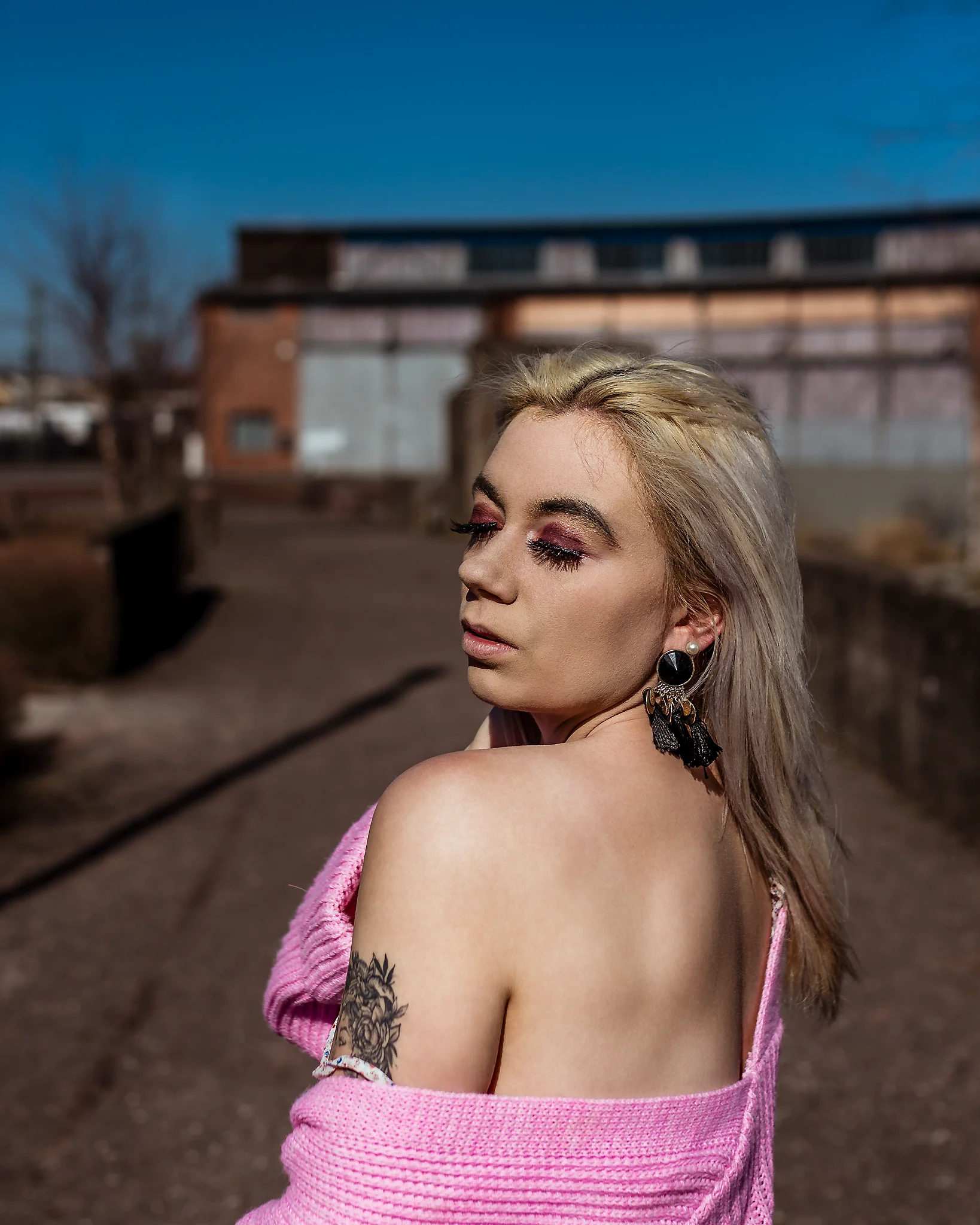 @chibiusa_luna_katie City - Person in urbaner Industrie-Umgebung mit freier Schulter und locker fallender, rosa Strickjacke, leicht über die Schulter blickend mit geschlossenen Augen und ruhigem, sinnlichem Ausdruck. Das harte Sonnenlicht modelliert Gesicht und Haut mit klaren Schatten, während der unscharfe Hintergrund aus Backstein und Metall eine reduzierte, moderne Bildtiefe erzeugt und den Fokus gezielt auf die Person lenkt.