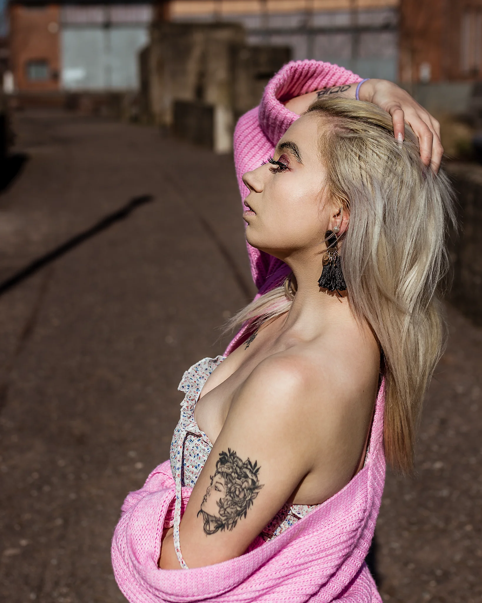 @chibiusa_luna_katie City - Person im seitlichen Profil in urbaner Industrie-Umgebung, mit angehobenem Arm im Haar und locker über die Schultern fallender, rosa Strickjacke, die eine natürliche, sinnliche Pose betont. Das harte Sonnenlicht hebt Hautstruktur, Make-up und Konturen deutlich hervor, während der weich verschwommene Hintergrund aus Backstein und Industrieelementen eine klare Trennung schafft und den Blick konsequent auf die Person lenkt.