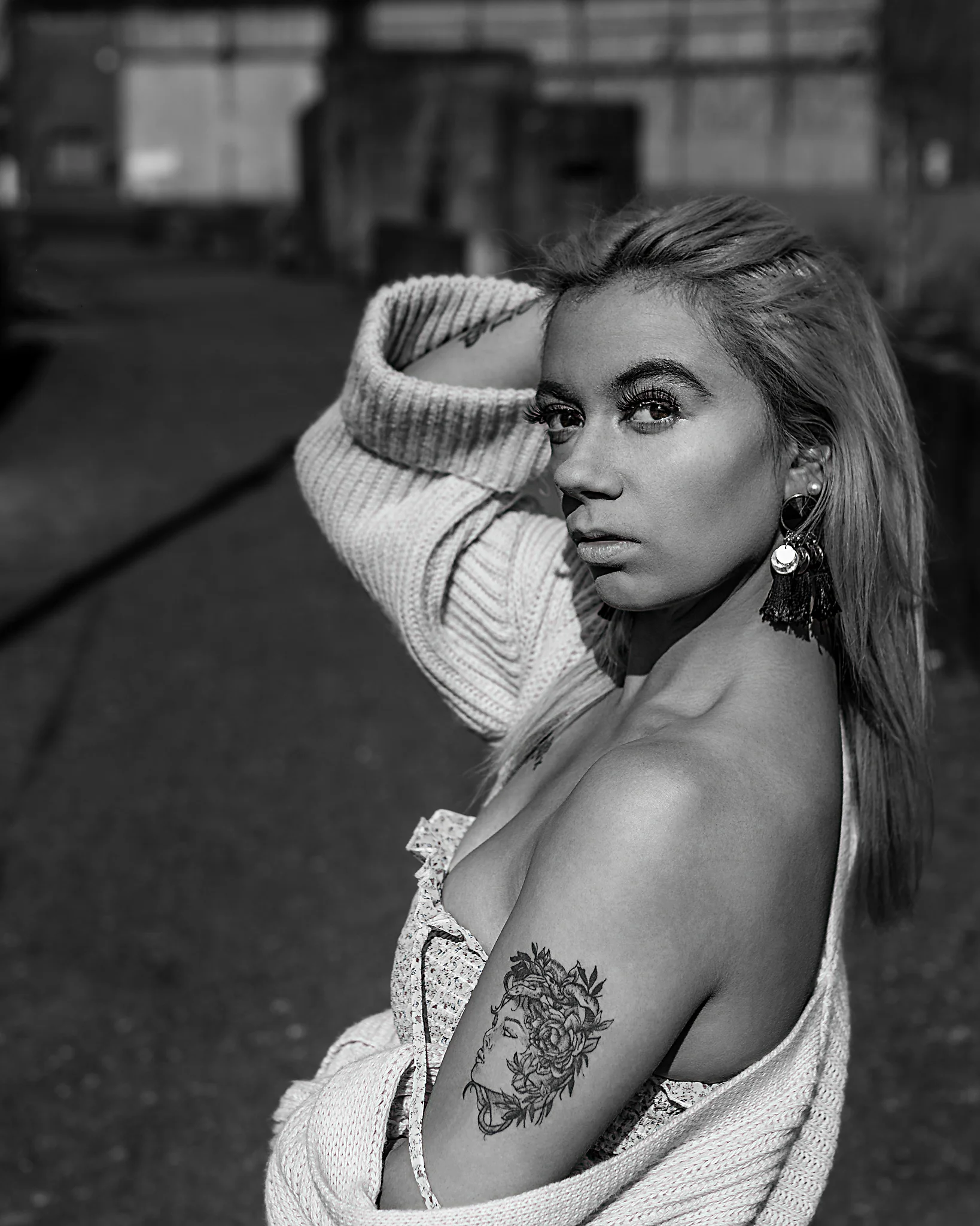 @chibiusa_luna_katie City - Person in urbaner Industrie-Umgebung in Schwarz-Weiß, mit angehobenem Arm im Haar und direktem, intensivem Blick zur Kamera, während die locker fallende Strickjacke die Schulter freilegt. Das harte Licht erzeugt starke Kontraste und betont Hautstruktur, Blick und Tattoo, während der unscharfe Hintergrund die Bildtiefe reduziert und den Fokus klar auf die Person lenkt.
