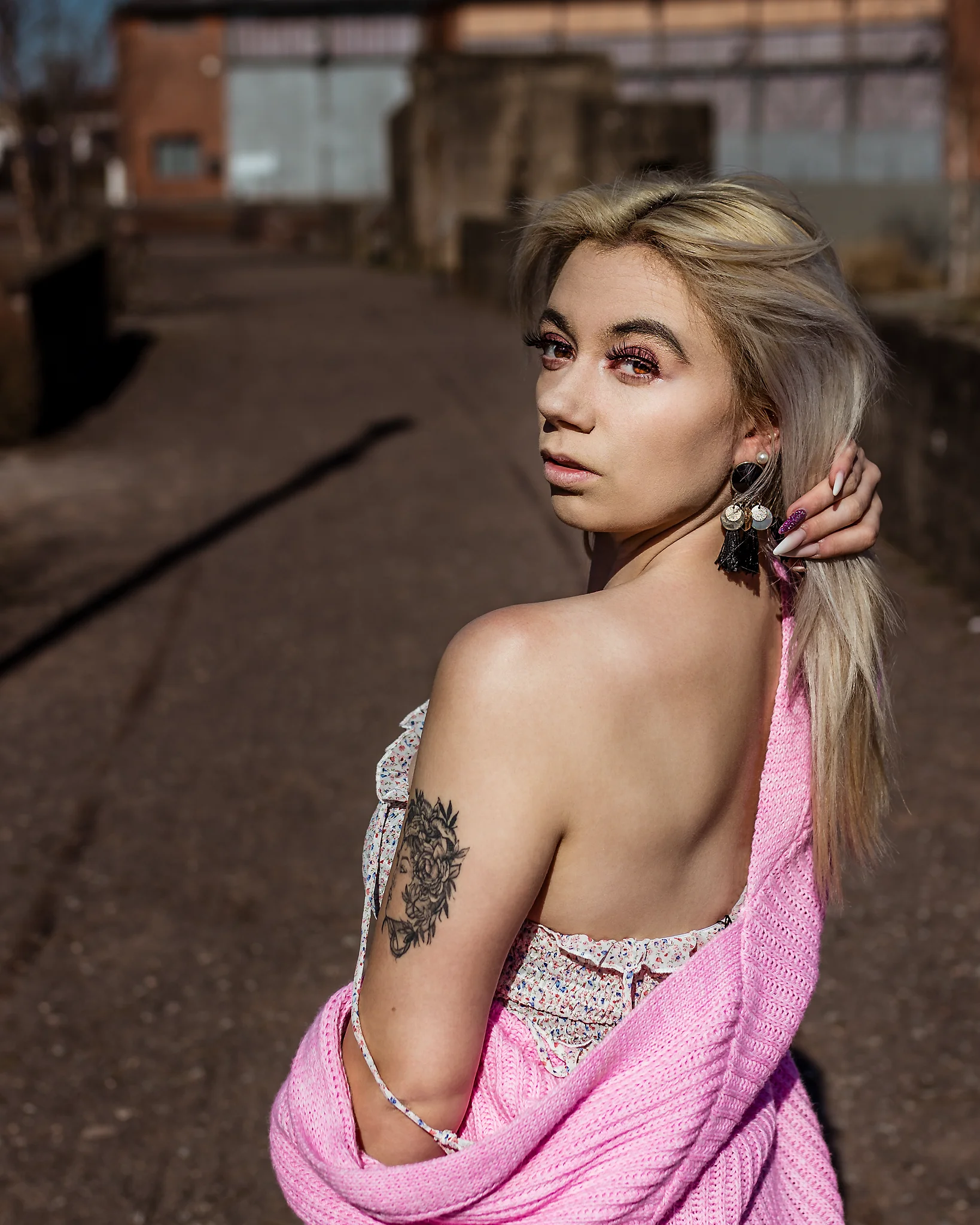 @chibiusa_luna_katie City - Person in urbaner Industrie-Umgebung, über die Schulter zur Kamera blickend mit direktem, ruhigem Ausdruck, während die locker fallende rosa Strickjacke die Rückenlinie betont. Das harte Sonnenlicht setzt klare Highlights auf Haut, Haare und Make-up, während der unscharfe Hintergrund aus Backstein und Industrieelementen eine reduzierte Tiefe schafft und den Fokus gezielt auf die Person lenkt.