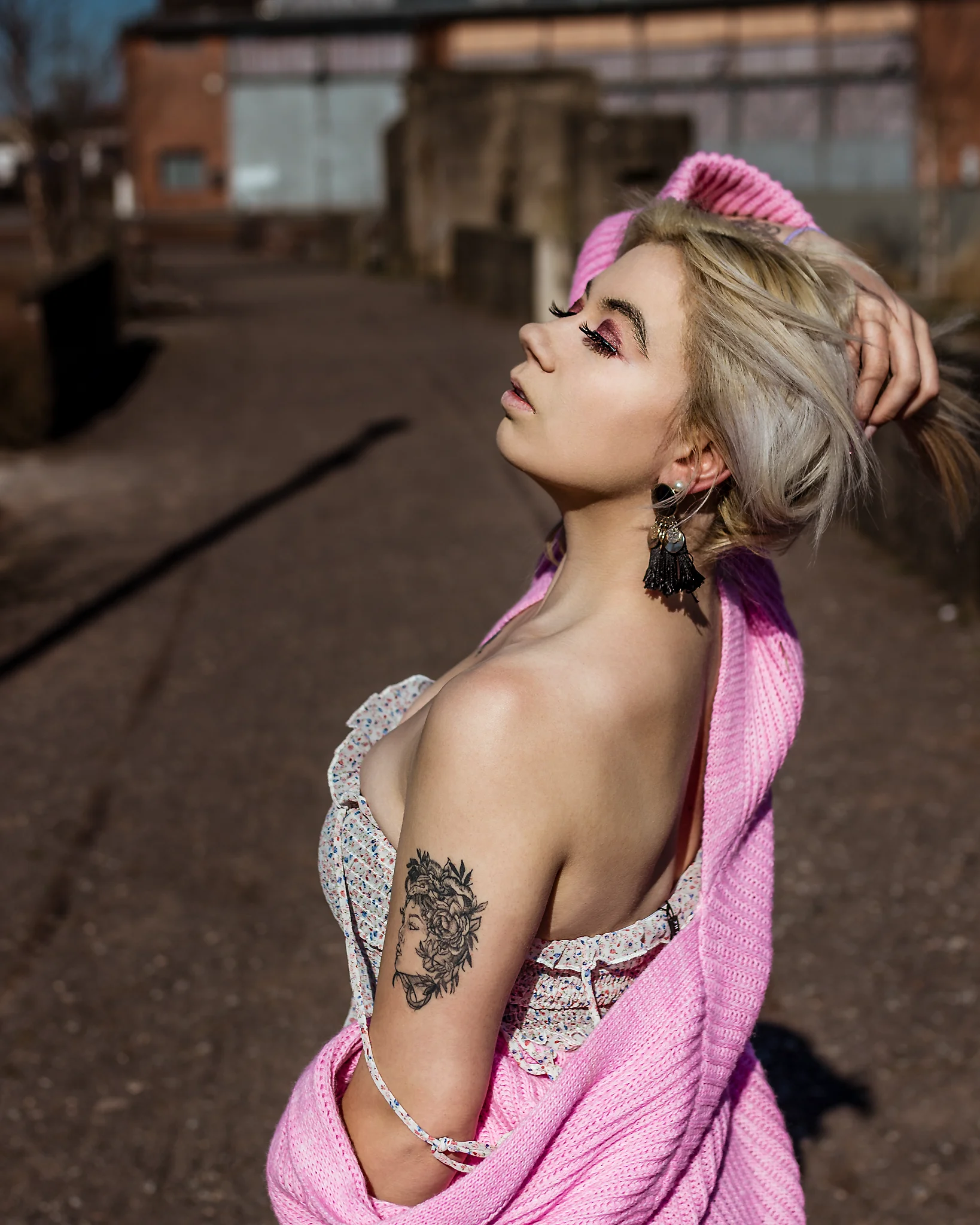 @chibiusa_luna_katie City - Person im seitlichen Profil in urbaner Industrie-Umgebung, mit nach hinten geneigtem Kopf und einer Hand im Haar, während die locker fallende rosa Strickjacke die Schulterpartie betont. Das harte Sonnenlicht erzeugt starke Kontraste und hebt Haut, Make-up sowie Konturen klar hervor, während der unscharfe Hintergrund aus Backstein und Industrieelementen eine reduzierte Tiefe schafft und den Fokus konsequent auf die Person lenkt.