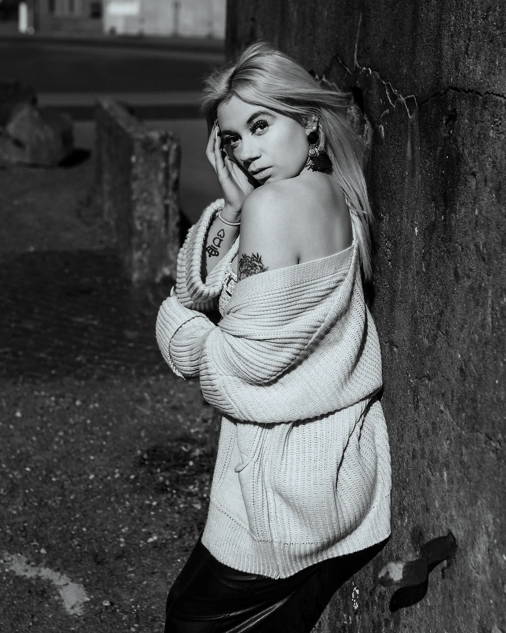 @chibiusa_luna_katie City - Person in urbaner Industrie-Umgebung in Schwarz-Weiß, an eine raue Betonwand gelehnt, mit leicht gedrehter Körperhaltung und ruhigem, intensiven Blick zur Kamera. Die locker fallende Strickjacke betont die Schulterpartie, während das gerichtete Licht starke Kontraste erzeugt und Struktur von Haut, Stoff und Wand hervorhebt, wodurch eine klare, reduzierte und atmosphärische Bildwirkung entsteht.