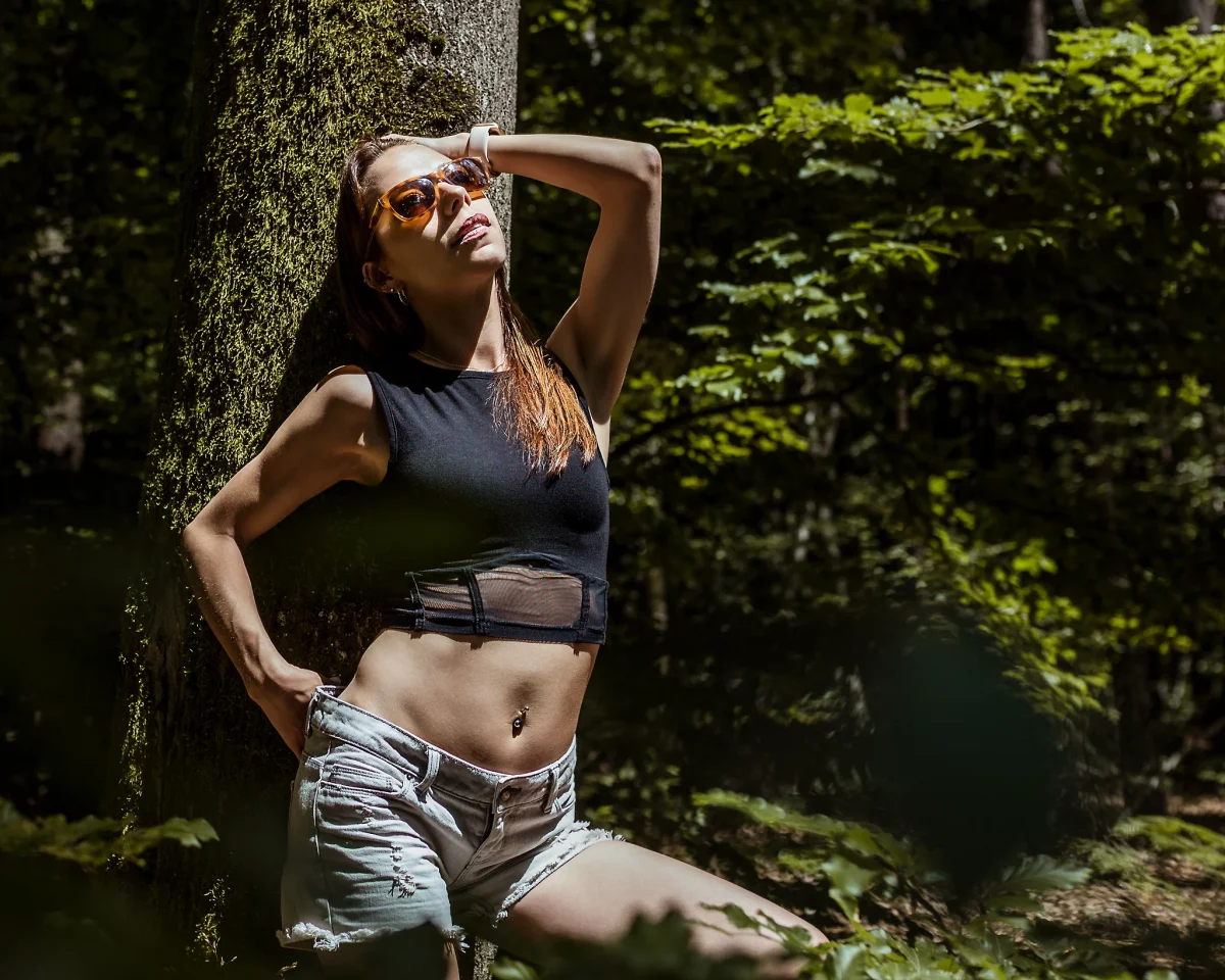 Natur - Person lehnt an einem Baum im Wald, trägt ein schwarzes Crop-Top und helle Jeansshorts, eine Sonnenbrille und hebt den Kopf ins Sonnenlicht, während eine Hand im Haar liegt; kontrastreiche Aufnahme mit hartem Licht und natürlichen Schatten im grünen Umfeld.