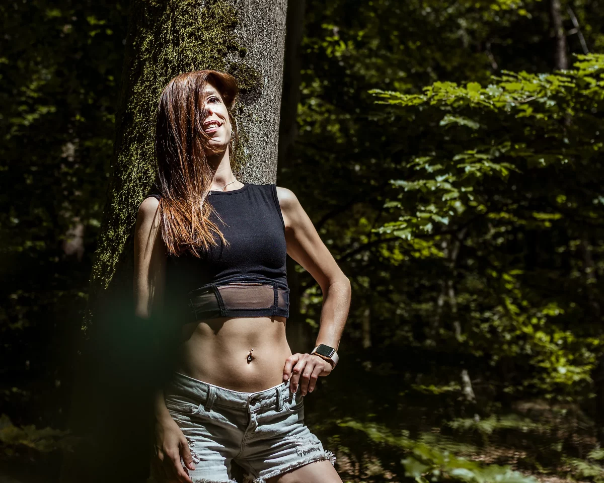 Natur - Person lehnt an einem moosbewachsenen Baum im Wald, trägt ein schwarzes Crop-Top und helle Jeansshorts, steht mit einer Hand in der Hüfte und blickt lächelnd ins Sonnenlicht; kontrastreiche Aufnahme mit starkem Licht-Schatten-Spiel im grünen Umfeld.