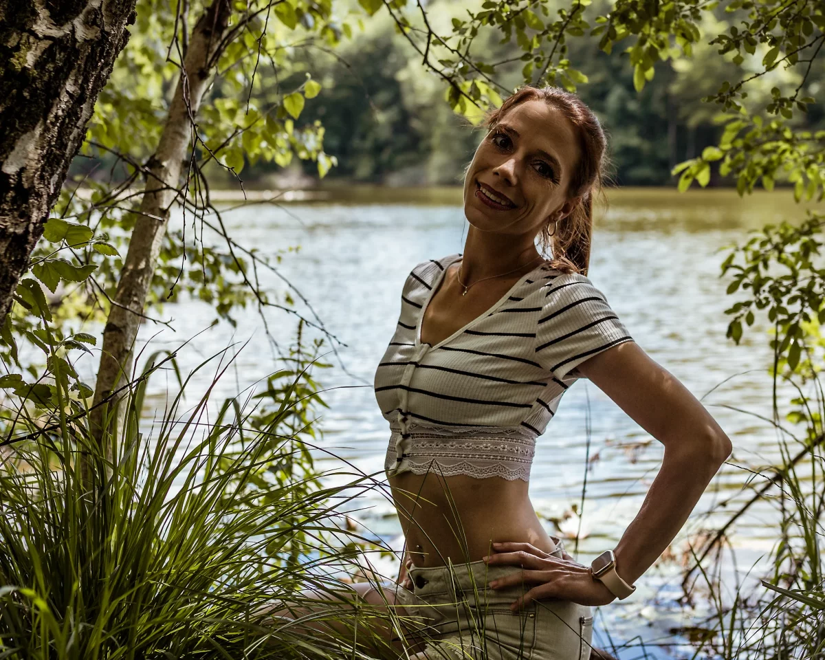 Natur - Person steht am Ufer eines Sees zwischen Gräsern und Bäumen, trägt ein gestreiftes Crop-Top und helle Shorts, stützt die Hände in die Hüfte und blickt lächelnd in die Kamera; natürliche Outdooraufnahme mit weichem Licht und ruhigem Wasser im Hintergrund.
