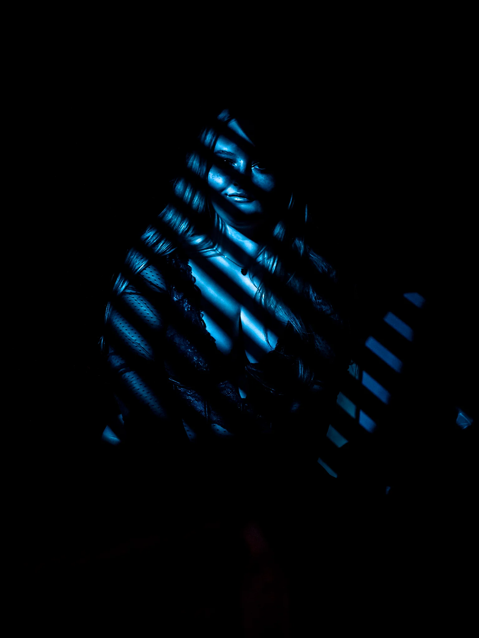 Creative - Dunkles, kontrastreiches Porträt einer Frau, die in nahezu vollständiger Schwärze steht, während schmale, gerichtete Lichtstreifen ihren Körper und ihr Gesicht in kühlen Blautönen zeichnen. Die Schattenstruktur erinnert an Jalousien oder Lamellen und erzeugt ein grafisches Spiel aus Licht und Dunkel, das nur Teile ihres Ausdrucks und ihrer Haltung sichtbar macht und eine geheimnisvolle, fast filmische Atmosphäre schafft.