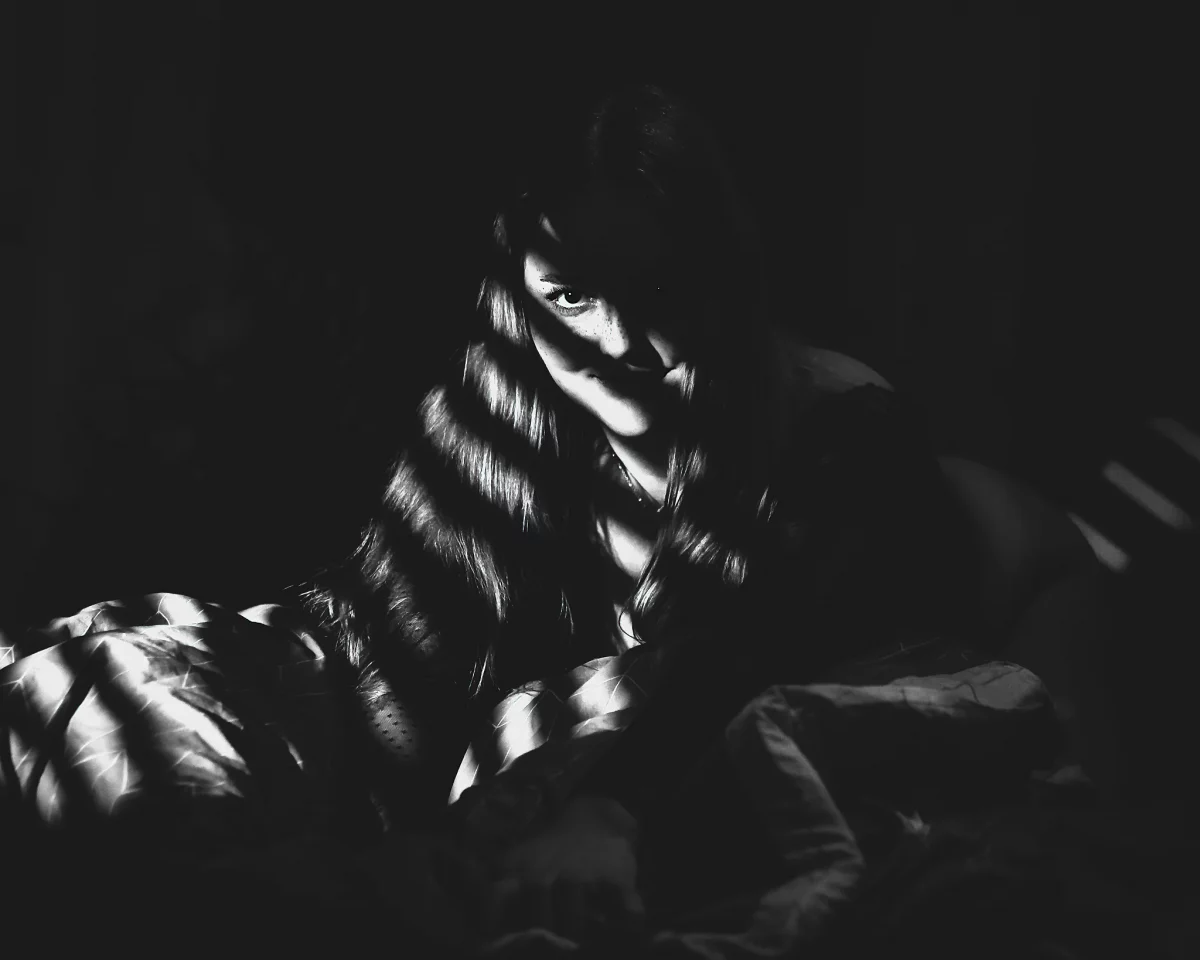 Creative - Kontrastreiches Schwarzweißporträt einer Frau, die im Dunkeln sitzt, teilweise in Decken gehüllt, während schmale Lichtstreifen ihr Gesicht und Haar durchziehen. Ihr direkter Blick aus dem Schatten heraus wirkt intensiv und leicht geheimnisvoll, während das harte Licht die Texturen von Stoff und Haar betont und eine dichte, filmische Atmosphäre erzeugt.