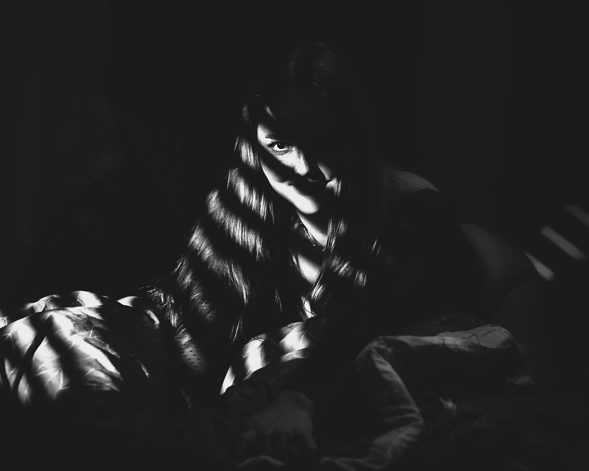 Creative - Kontrastreiches Schwarzweißporträt einer Frau, die im Dunkeln sitzt, teilweise in Decken gehüllt, während schmale Lichtstreifen ihr Gesicht und Haar durchziehen. Ihr direkter Blick aus dem Schatten heraus wirkt intensiv und leicht geheimnisvoll, während das harte Licht die Texturen von Stoff und Haar betont und eine dichte, filmische Atmosphäre erzeugt.