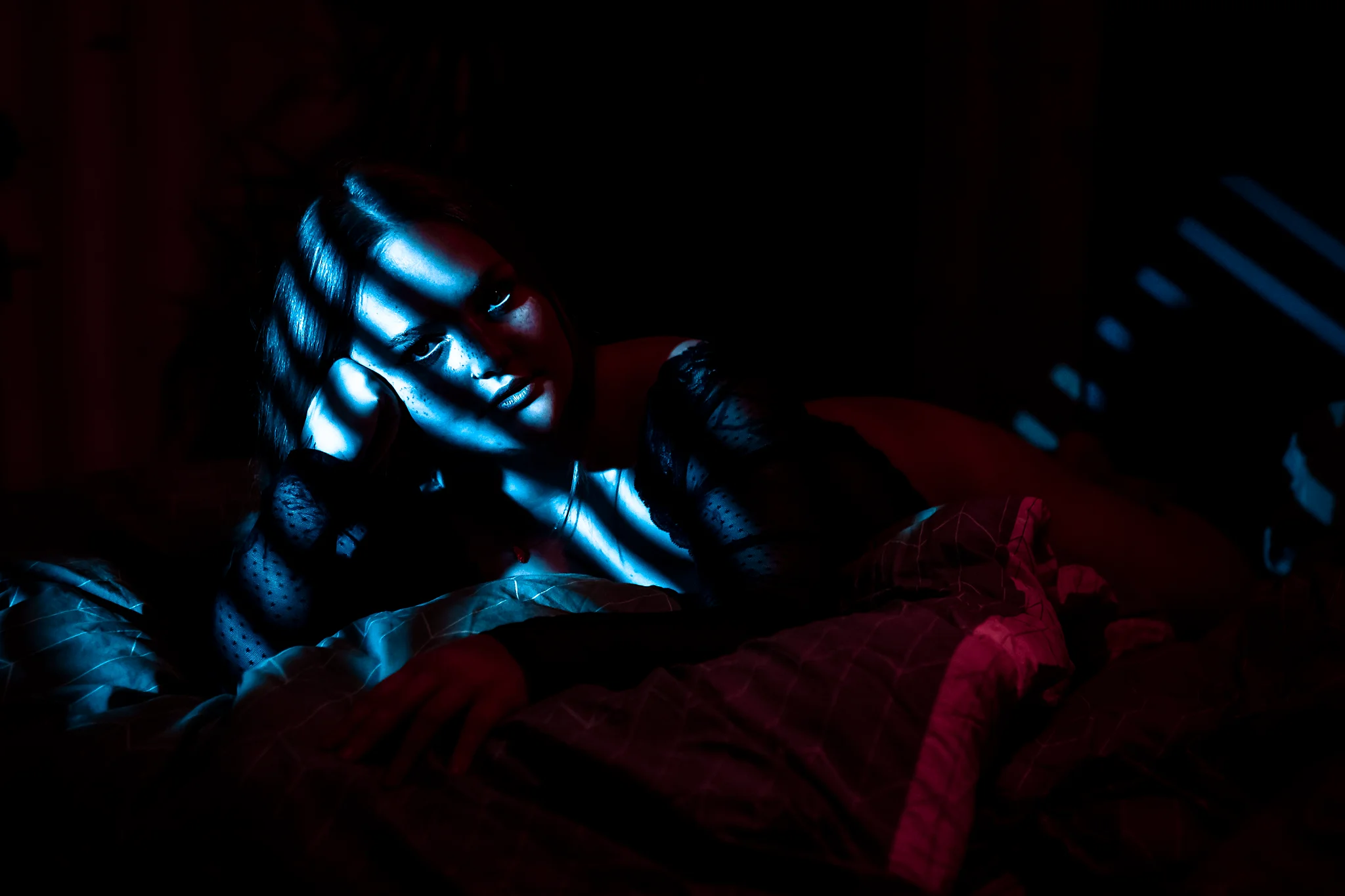 Creative - Stimmungsvolles Porträt einer Frau in dunkler Umgebung, die auf einem Bett liegt und ihren Kopf in die Hand stützt, während schmale, harte Lichtstreifen in kühlen Blautönen ihr Gesicht und ihren Oberkörper strukturieren. Ergänzt wird das Licht durch warme, rötliche Schatten im Umfeld, wodurch ein spannungsreicher Farbkontrast entsteht. Die Mischung aus Licht und Dunkelheit verleiht dem Bild eine intime, leicht geheimnisvolle und cineastische Atmosphäre.