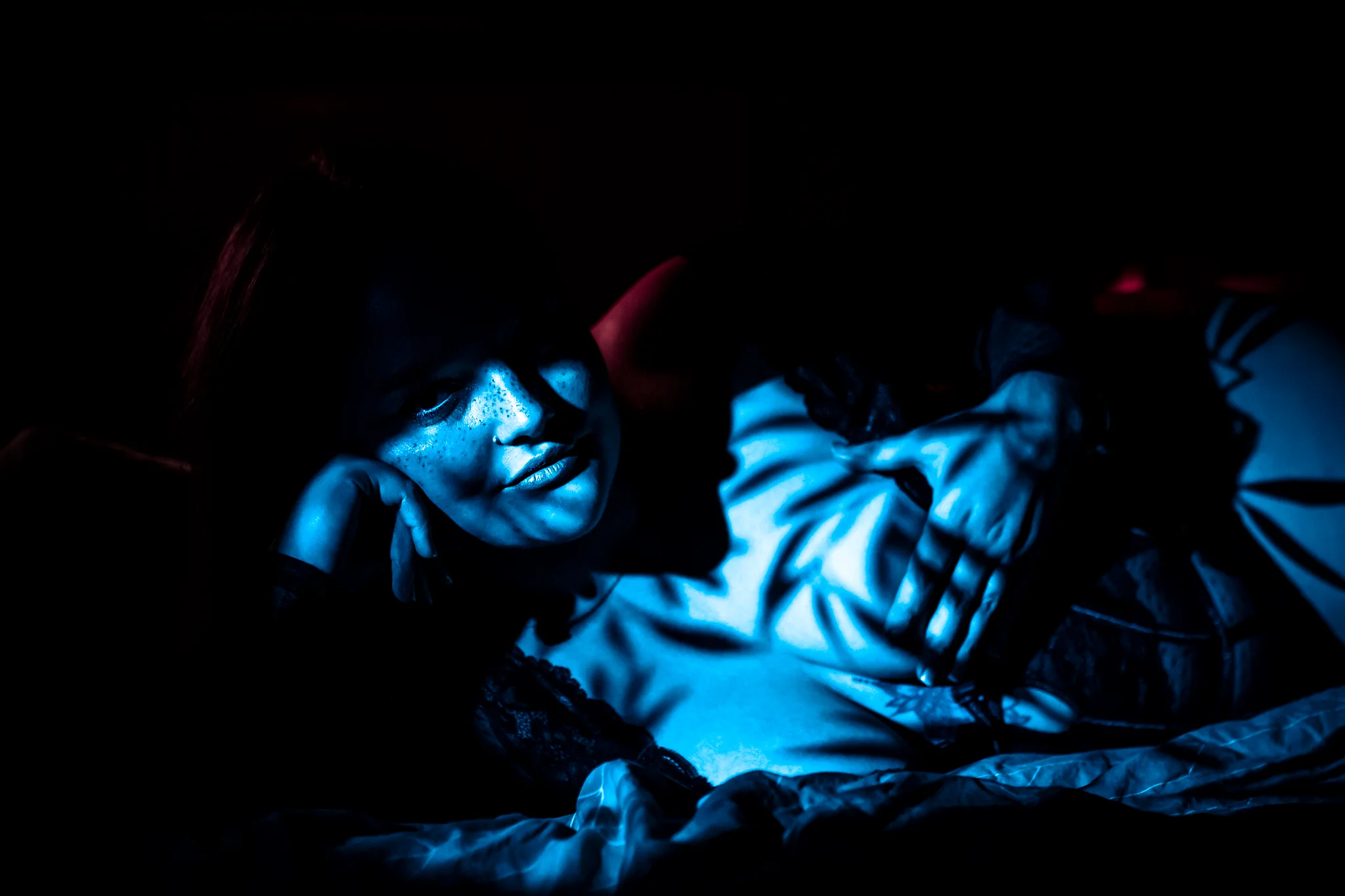Creative - Atmosphärisches Porträt einer Frau in dunkler, cineastischer Lichtstimmung, die liegend inszeniert ist und ihren Kopf auf die Hand stützt, während ihr Körper leicht eingedreht wirkt. Kühles, gerichtetes Licht in intensiven Blautönen modelliert Gesicht und Oberkörper, während warme rötliche Akzente im Hintergrund für Kontrast sorgen. Das Spiel aus Lichtstreifen und Schatten verdeckt Teile ihres Gesichts und erzeugt eine sinnliche, geheimnisvolle und zugleich ruhige Bildwirkung.