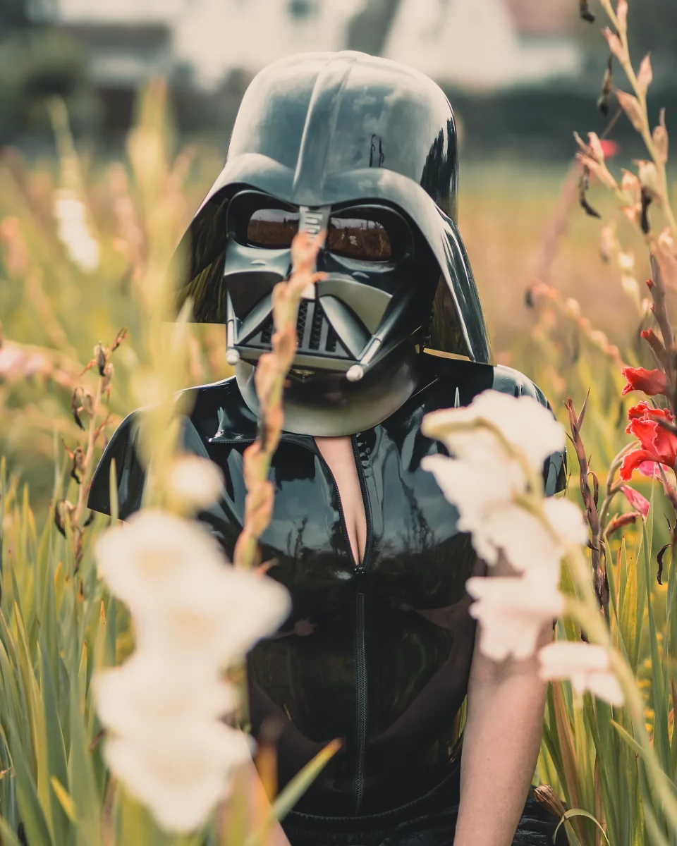 Star Wars girl's - Person in einem schwarzen Latex-Outfit trägt einen Darth-Vader-Helm und steht zwischen hohen Blumen auf einer Wiese, wodurch ein surrealer Kontrast zwischen Natur und ikonischer Sci-Fi-Ästhetik entsteht.