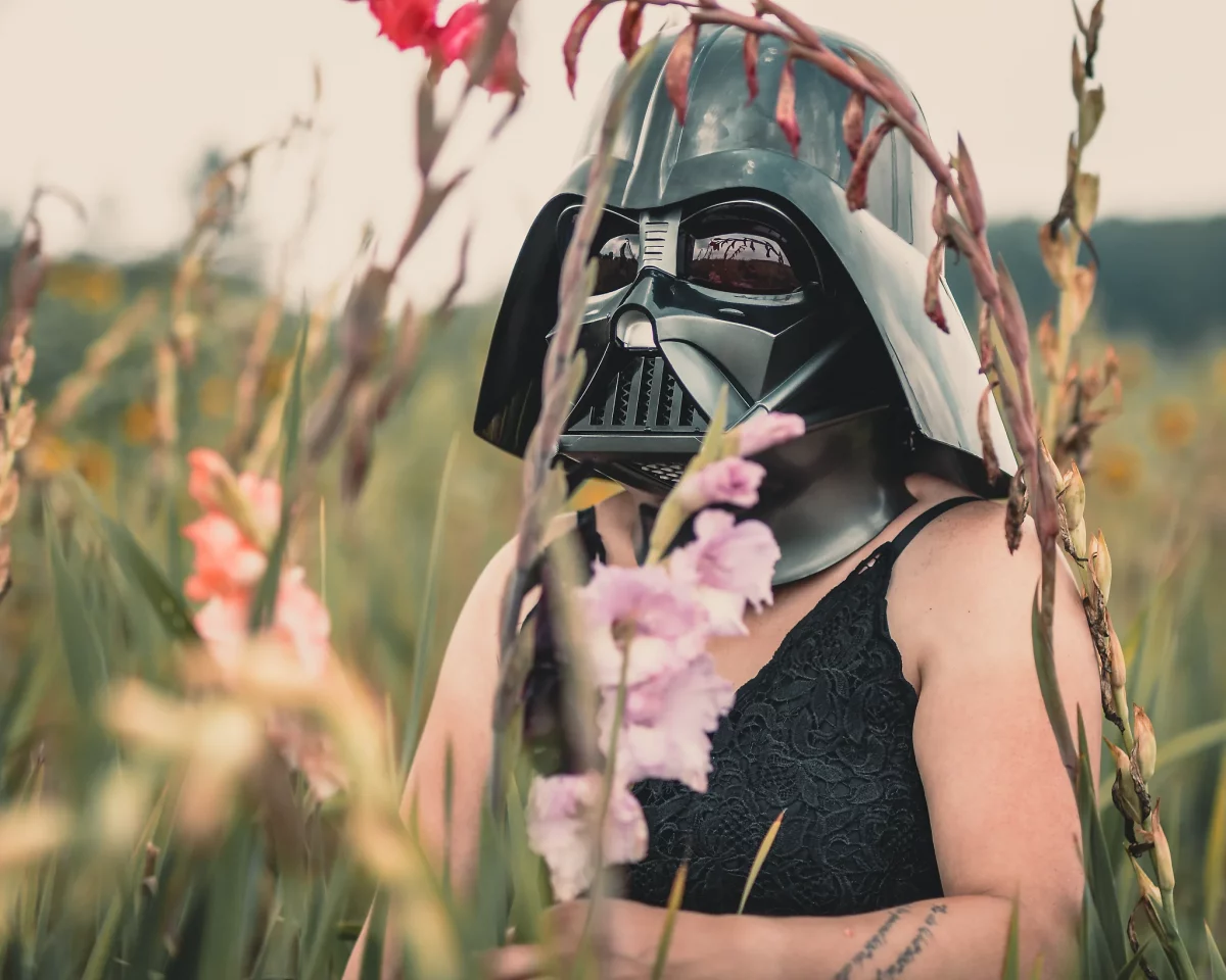 Star Wars girl's - Person mit schwarzem Spitzen-Top trägt einen Darth-Vader-Helm und steht zwischen blühenden Pflanzen auf einer Wiese, wodurch ein kontrastreicher Mix aus Natur und ikonischer Sci-Fi-Ästhetik entsteht.
