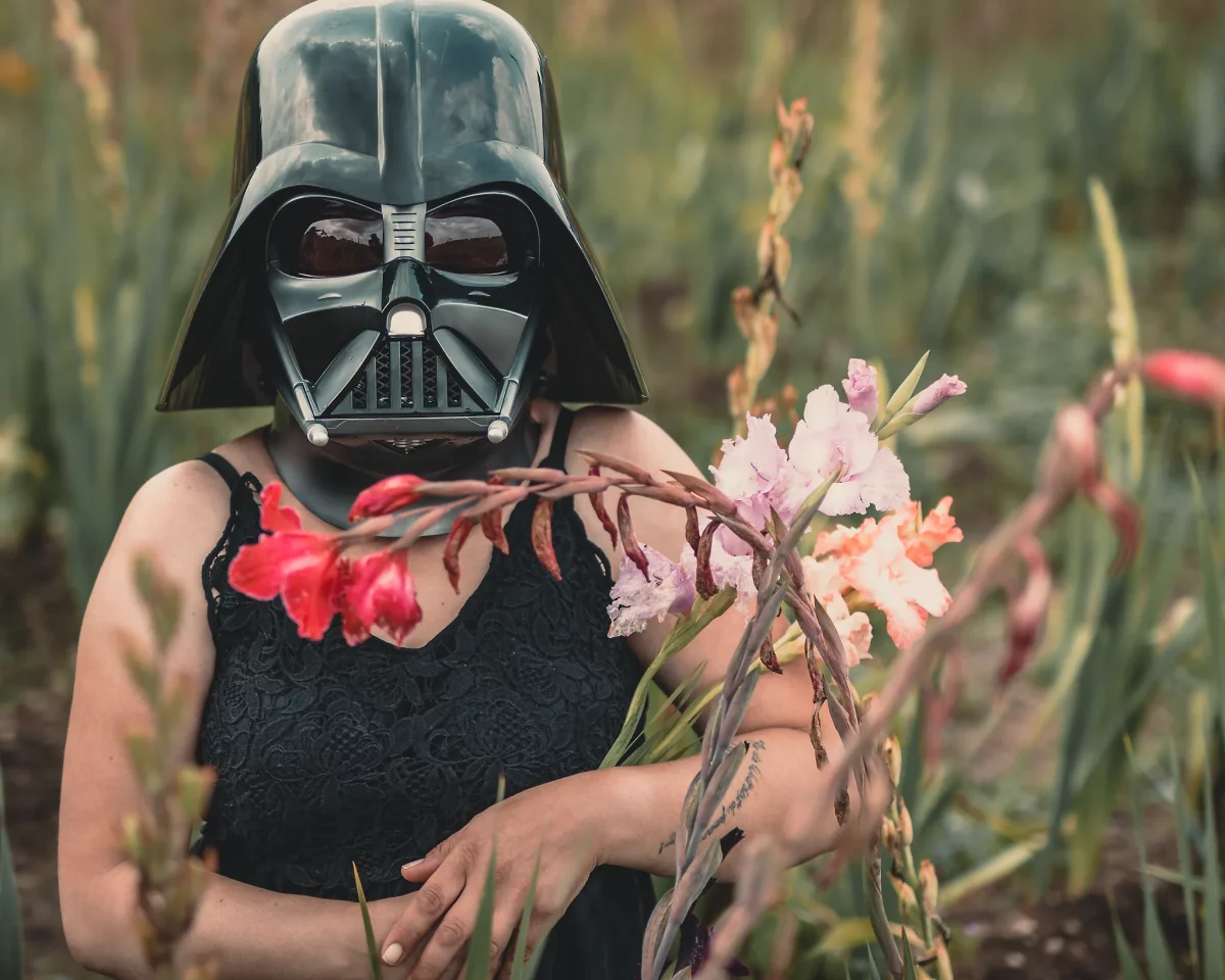 Star Wars girl's - Person mit schwarzem Spitzen-Top trägt einen Darth-Vader-Helm und sitzt zwischen farbigen Blüten auf einer Wiese, wodurch ein surrealer Kontrast zwischen Natur und Sci-Fi-Ästhetik entsteht.