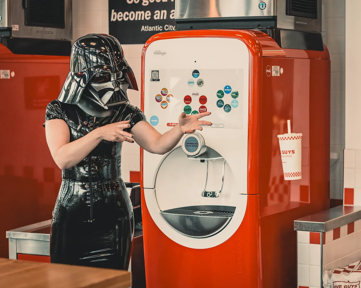 Star Wars girl's - Person im schwarzen Latex-Outfit trägt einen Darth-Vader-Helm und steht neben einem Getränkeautomaten in einem Fast-Food-Restaurant, während sie mit einer „Macht“-Geste einen Becher schweben lässt.