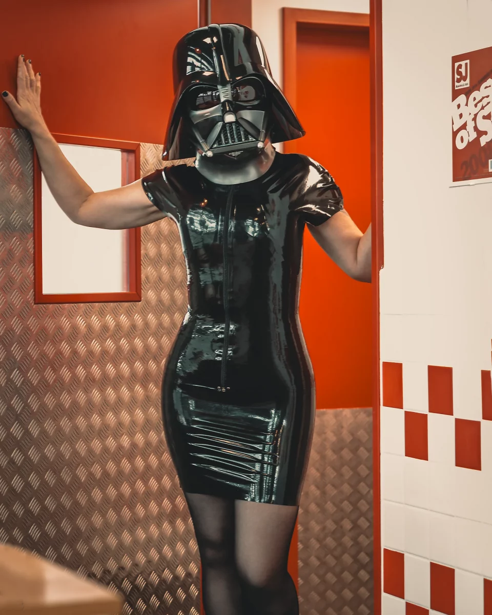 Star Wars girl's - Person im schwarzen Latexkleid trägt einen Darth-Vader-Helm und steht in einem rot-weiß gestalteten Innenraum in einer Türöffnung, wobei die glänzende Oberfläche des Outfits und die klare Farbgebung eine starke, grafische Bildwirkung erzeugen.