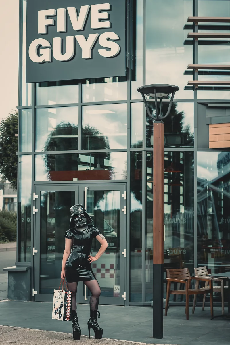 Star Wars girl's - Person im schwarzen Latexkleid und High Heels trägt einen Darth-Vader-Helm und posiert selbstbewusst vor dem Eingang eines Five Guys Restaurants, während sie eine Einkaufstasche in der Hand hält.