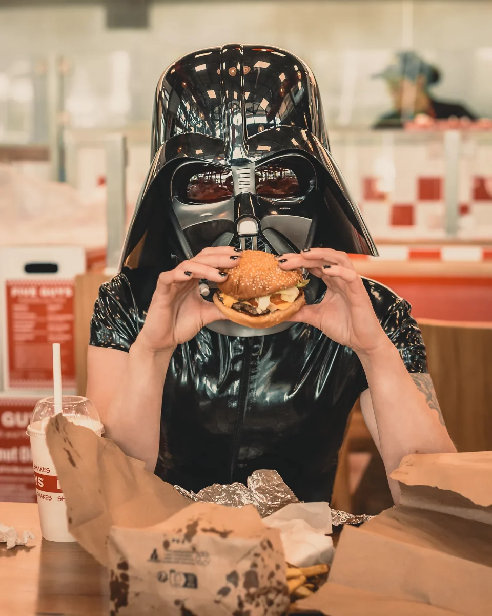 Star Wars girl's - Person im schwarzen Latex-Outfit trägt einen Darth-Vader-Helm und hält einen Burger vor sich, während sie an einem Tisch in einem Fast-Food-Restaurant sitzt.