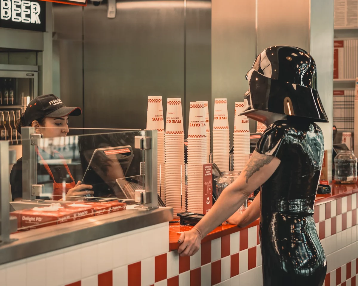 Star Wars girl's - Person im schwarzen Latex-Outfit trägt einen Darth-Vader-Helm und steht an der Theke eines Fast-Food-Restaurants, während sie eine Bestellung aufgibt.