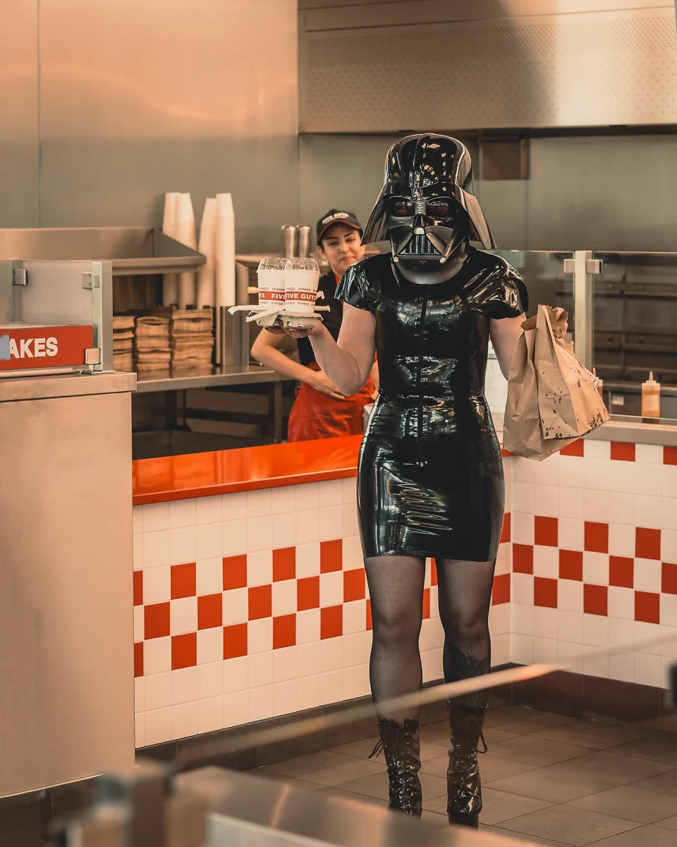Star Wars girl's - Person im schwarzen Latexkleid trägt einen Darth-Vader-Helm und verlässt den Tresen eines Fast-Food-Restaurants mit Getränken und einer Tüte in der Hand.