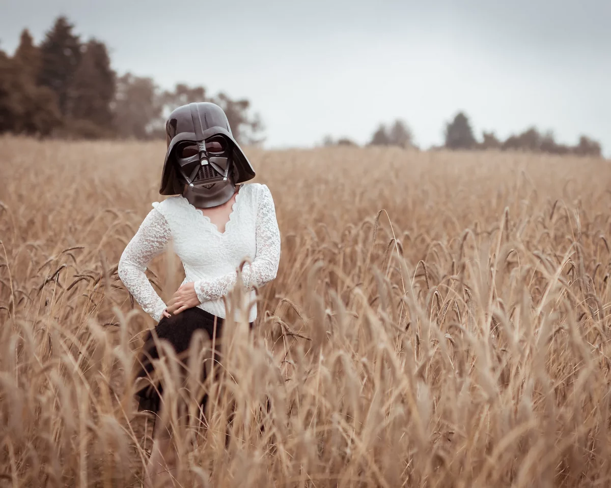 Star Wars girl's - Person mit weißer Spitzenbluse und dunklem Rock trägt einen Darth-Vader-Helm und steht in einem weiten Getreidefeld, wobei die Pose ruhig und leicht versetzt wirkt.