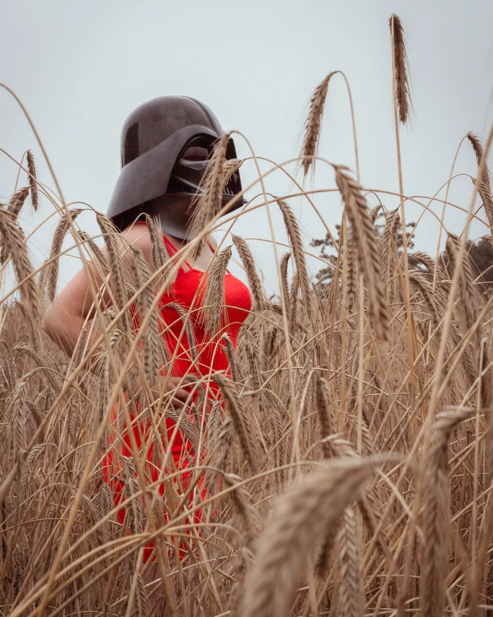 Star Wars girl's - Person im roten Kleid trägt einen Darth-Vader-Helm und steht zwischen hohen Getreidehalmen, die den Vordergrund dicht umrahmen.