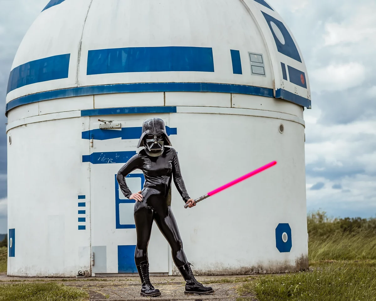 Star Wars girl's - Person im schwarzen Latex-Catsuit und mit Darth-Vader-Helm steht vor der R2D2-Sternwarte und hält ein leuchtendes rosa Lichtschwert in der Hand.