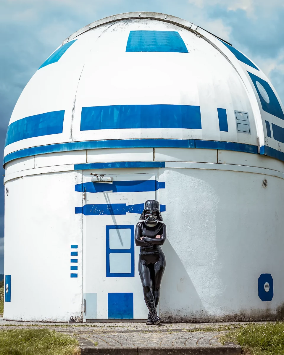 Star Wars girl's - Person im schwarzen Latex-Catsuit und mit Darth-Vader-Helm lehnt mit verschränkten Armen vor der R2D2-Sternwarte.