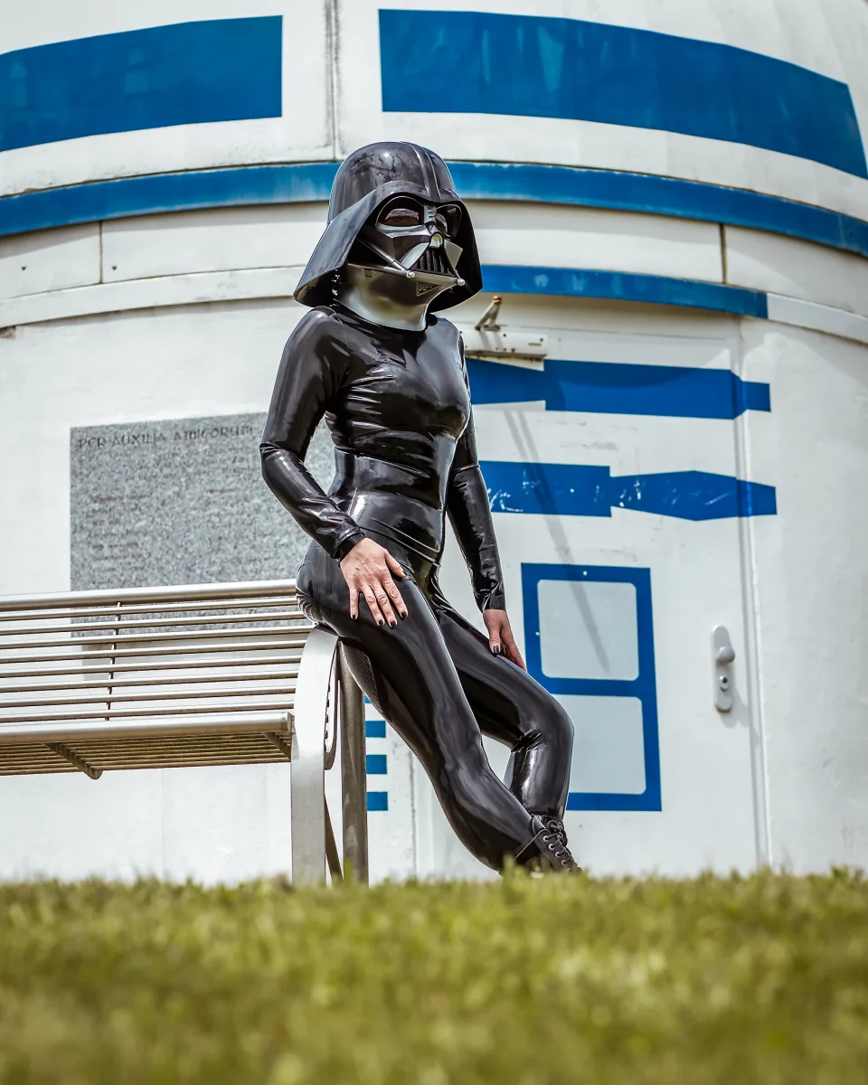 Star Wars girl's - Person im schwarzen Latex-Catsuit und mit Darth-Vader-Helm sitzt seitlich auf einer Metallbank vor der R2D2-Sternwarte.