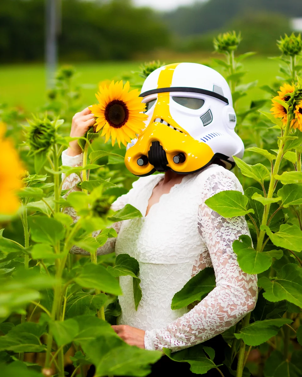 Star Wars girl's - Person in einem weißen Spitzenbody trägt einen gelb-weißen Stormtrooper-Helm und hält eine Sonnenblume neben dem Kopf, umgeben von hohen Sonnenblumenpflanzen.