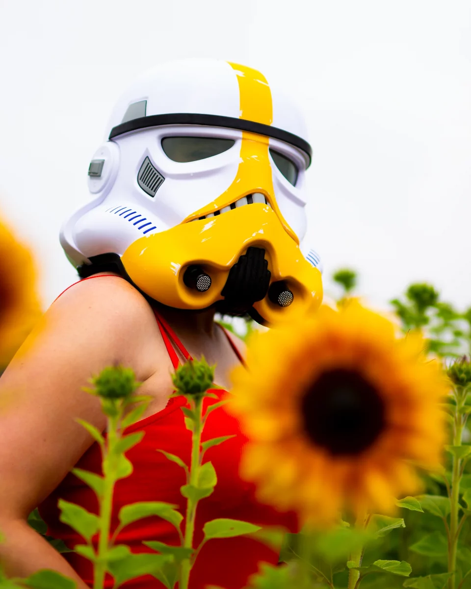 Star Wars girl's - Person im roten Body trägt einen gelb-weißen Stormtrooper-Helm und steht zwischen Sonnenblumen, eine Blüte unscharf im Vordergrund.