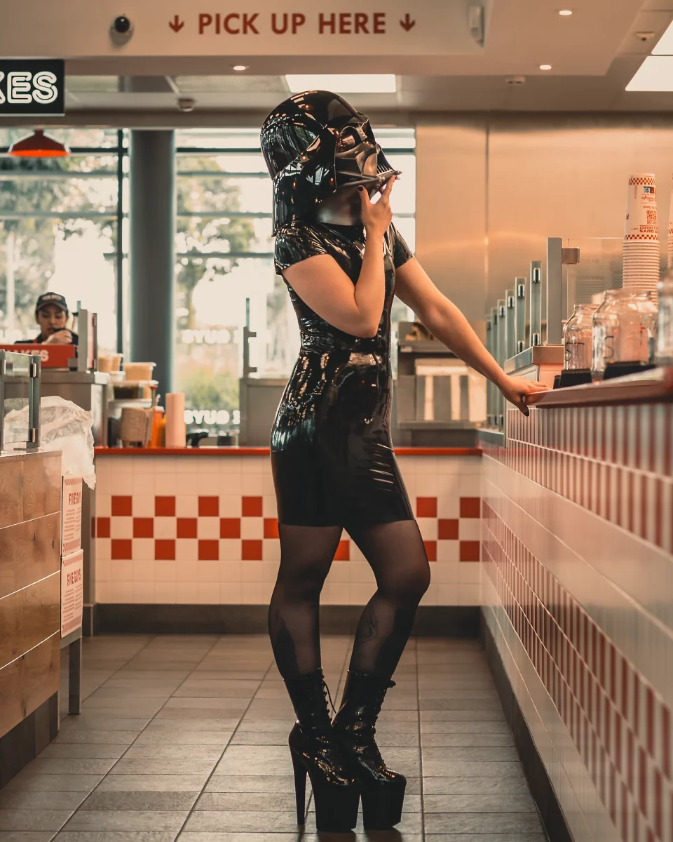 Star Wars girl's - Person im schwarzen Latexkleid und High Heels mit Darth-Vader-Helm steht am Tresen eines Fast-Food-Restaurants und berührt nachdenklich das Kinn, während im Hintergrund „Pick Up Here“ zu sehen ist.