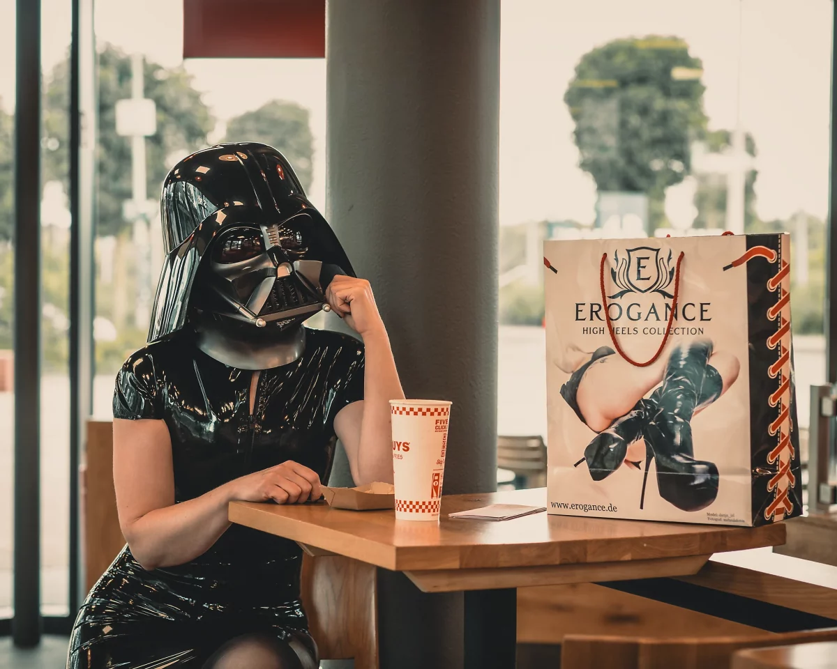 Star Wars girl's - Person im schwarzen Latex-Kleid und mit Darth-Vader-Helm sitzt an einem Tisch im Fast-Food-Restaurant, stützt den Kopf nachdenklich auf die Hand, während neben ihr eine Einkaufstasche und ein Getränk stehen.