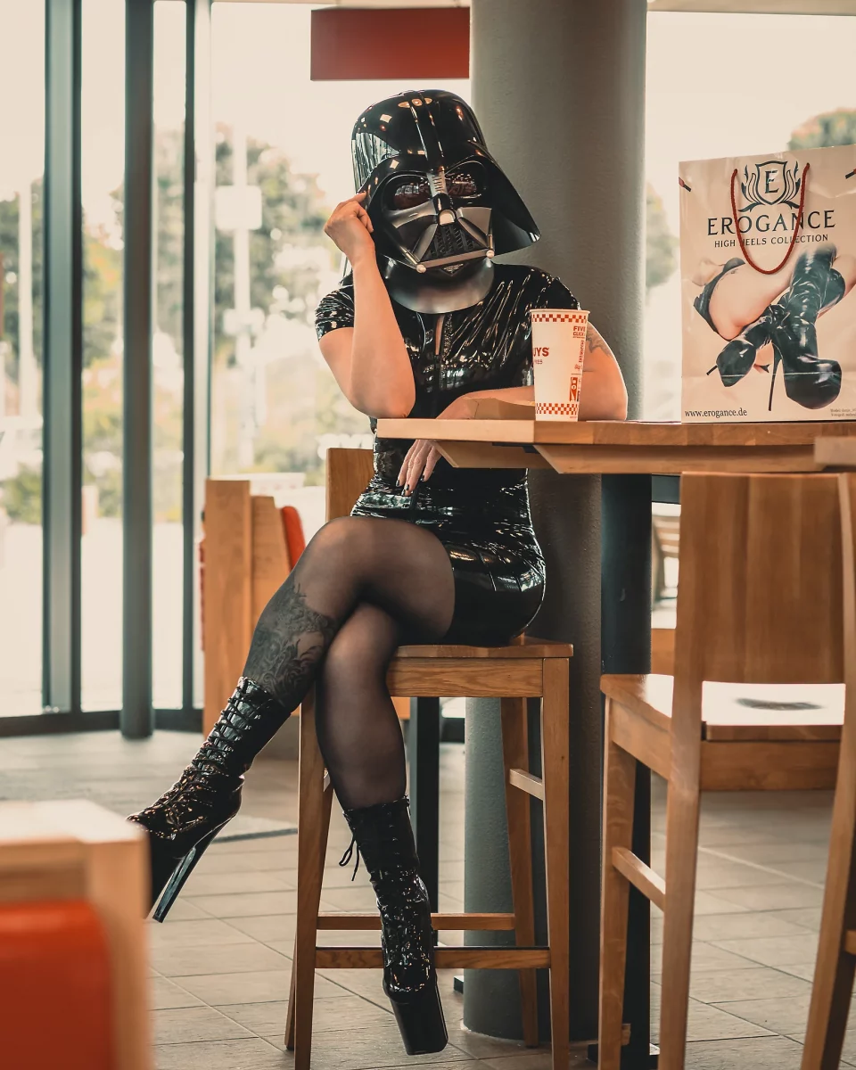 Star Wars girl's - Person im schwarzen Latex-Kleid und mit Darth-Vader-Helm sitzt auf einem hohen Stuhl im Fast-Food-Restaurant, hat die Beine übereinandergeschlagen und stützt den Kopf nachdenklich auf die Hand, während auf dem Tisch ein Getränk und eine Einkaufstasche stehen.