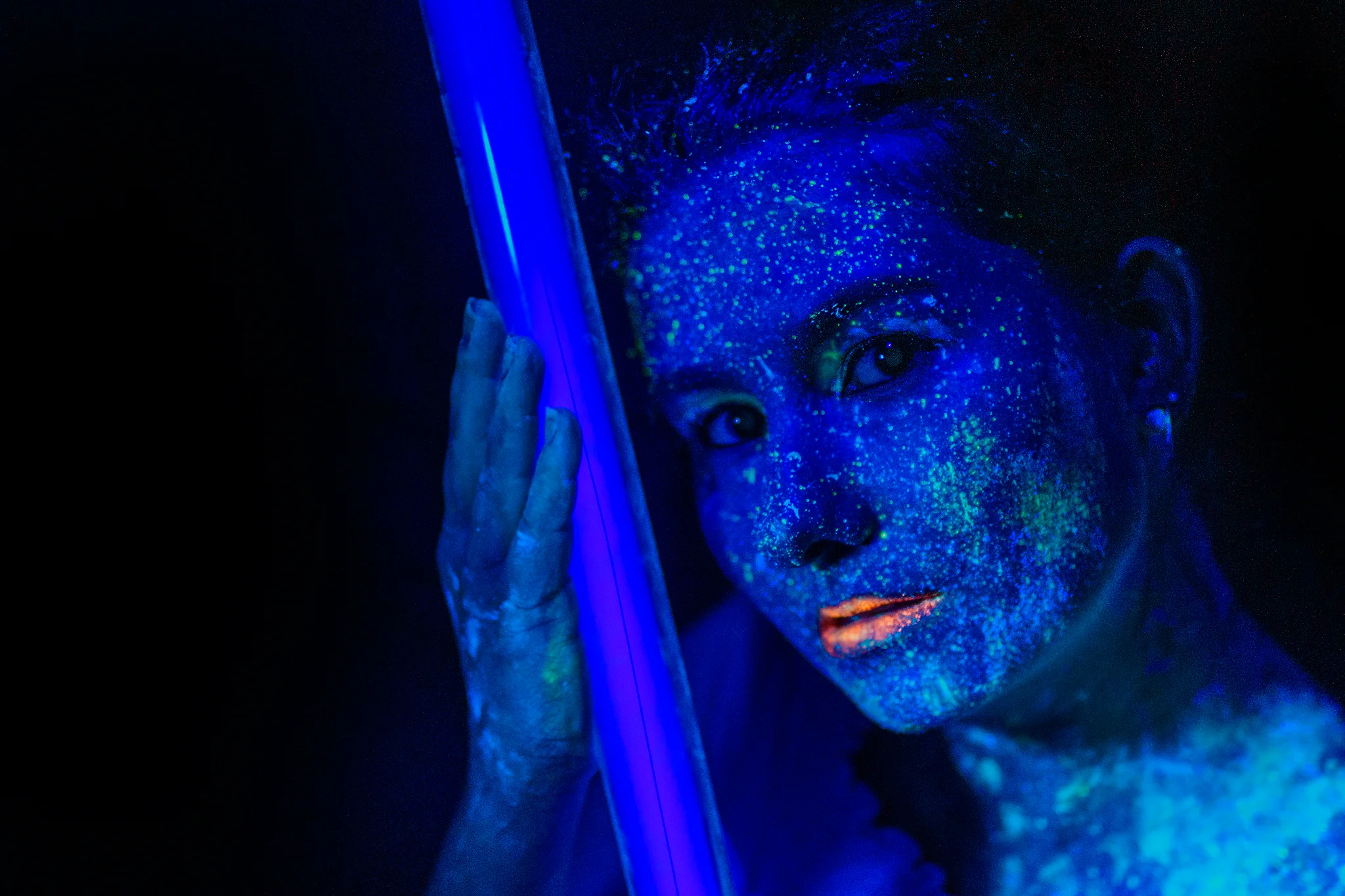 Creative - Intensives Porträt einer Frau in UV-Licht, deren Gesicht und Oberkörper mit fluoreszierenden Pigmenten bedeckt sind, die in leuchtenden Blau- und Grüntönen schimmern. Sie lehnt sich nah an eine vertikale Lichtquelle, die ihr Gesicht zusätzlich akzentuiert, während ihre Lippen in warmem Kontrast hervorstechen. Der dunkle Hintergrund verstärkt die Strahlkraft der Farben und verleiht dem Bild eine futuristische, fast außerweltliche Atmosphäre.