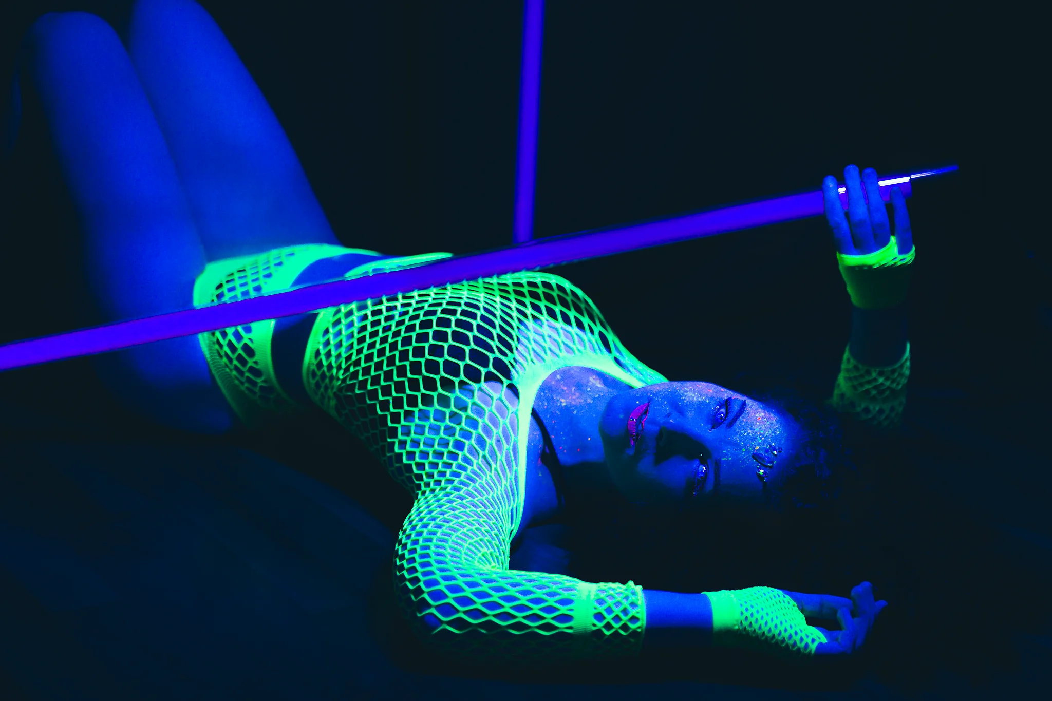Creative - Futuristisch wirkendes Porträt einer Frau im UV-Licht, die liegend inszeniert ist und einen leuchtenden Stab über ihrem Körper hält. Ihr neonfarbenes Netzoutfit fluoresziert intensiv in Grün, während Haut und Gesicht in kühlen Blautönen schimmern und mit feinen, glitzernden Pigmenten überzogen sind. Der dunkle Hintergrund verstärkt die Leuchtkraft der Farben und erzeugt eine stilisierte, fast außerweltliche Atmosphäre mit starkem grafischen Kontrast.