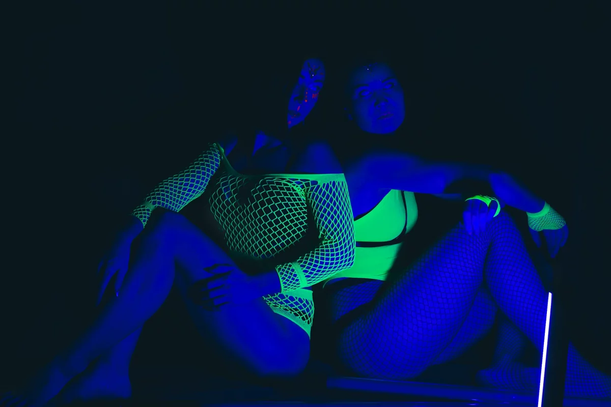 Zwei Frauen sitzen eng nebeneinander in einer dunklen UV-Lichtszene, ihre Haut erscheint blau, während neon-grüne Netzkleidung und ein leuchtender Stab intensiv fluoreszieren und eine futuristische Atmosphäre erzeugen.
