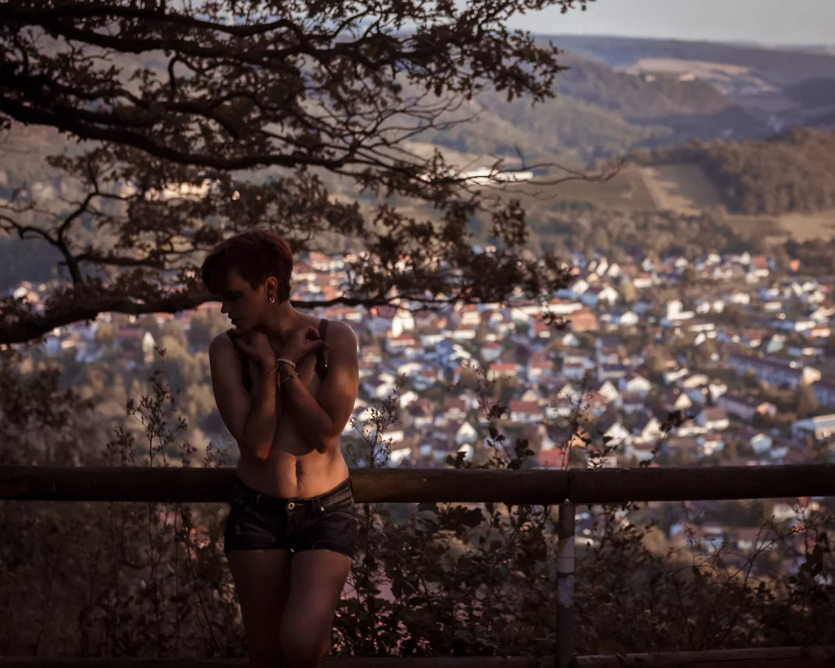 Natur - Person steht an einem Aussichtspunkt oberhalb einer Stadtlandschaft, umgeben von Bäumen und warmem Abendlicht. Die Pose wirkt ruhig und leicht verschlossen, mit seitlich abgewandtem Blick. Das weiche, goldene Licht modelliert den Körper dezent, während der unscharfe Hintergrund mit Häusern und Hügeln eine ruhige, leicht melancholische Atmosphäre erzeugt.