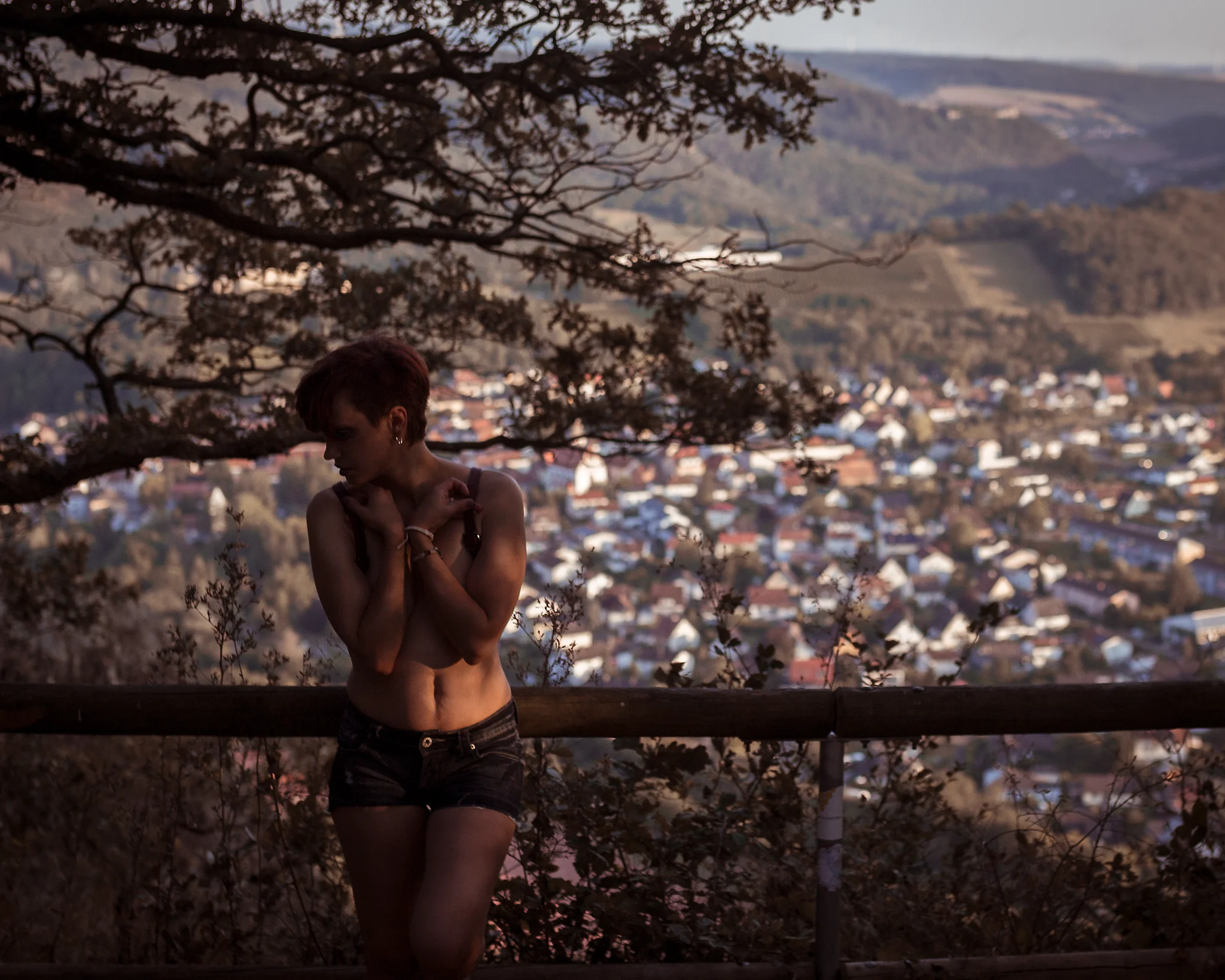 Natur - Person steht an einem Aussichtspunkt oberhalb einer Stadtlandschaft, umgeben von Bäumen und warmem Abendlicht. Die Pose wirkt ruhig und leicht verschlossen, mit seitlich abgewandtem Blick. Das weiche, goldene Licht modelliert den Körper dezent, während der unscharfe Hintergrund mit Häusern und Hügeln eine ruhige, leicht melancholische Atmosphäre erzeugt.